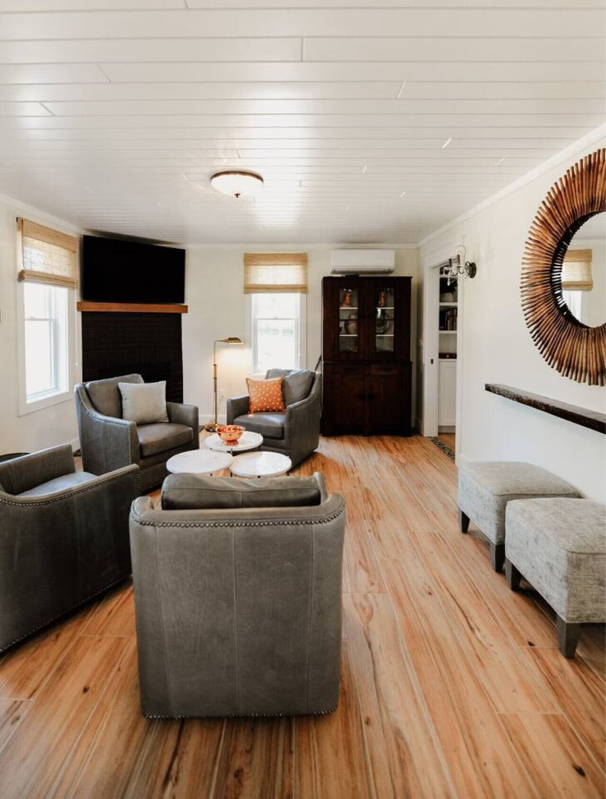 Living room with wooden flooring, gray armchairs, round white coffee tables, a dark wooden cabinet, large windows with blinds, a wall mirror, and a television mounted above a fireplace