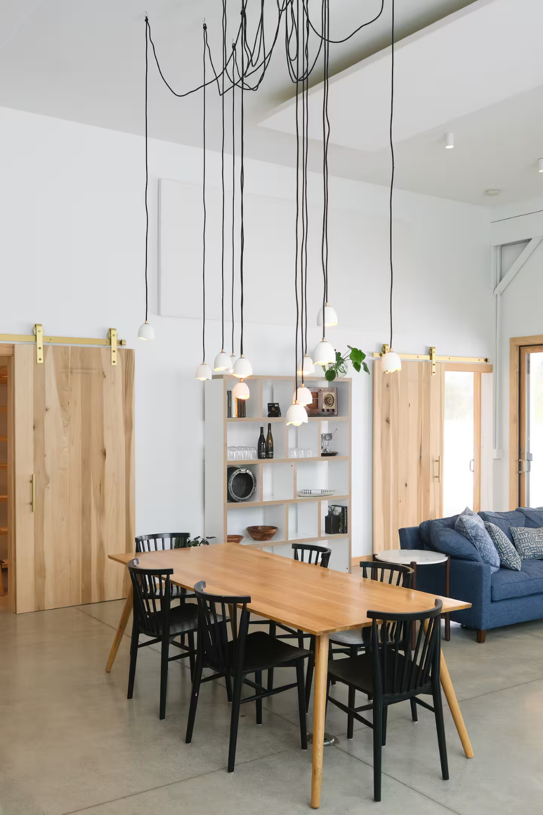 A modern dining and living room area with a wooden dining table and black chairs, a blue sofa with patterned pillows, a white bookshelf with decorative items, sliding wooden barn doors, a window, and hanging pendant lights from a black wire fixture.