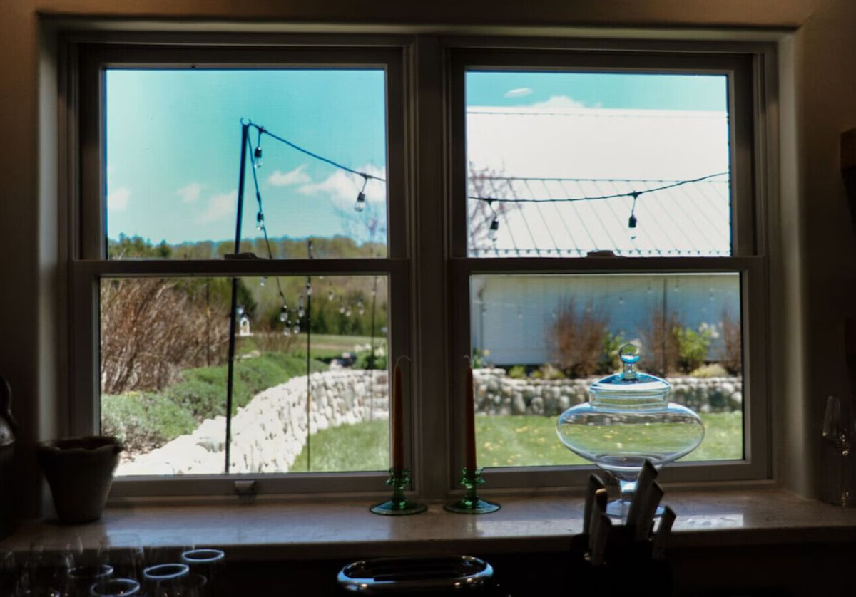 View of a bright, sunny outdoor scene through a four-pane window, with string lights hanging outside, trees, a stone wall, grass, and a building visible in the background, along with indoor objects on the windowsill.