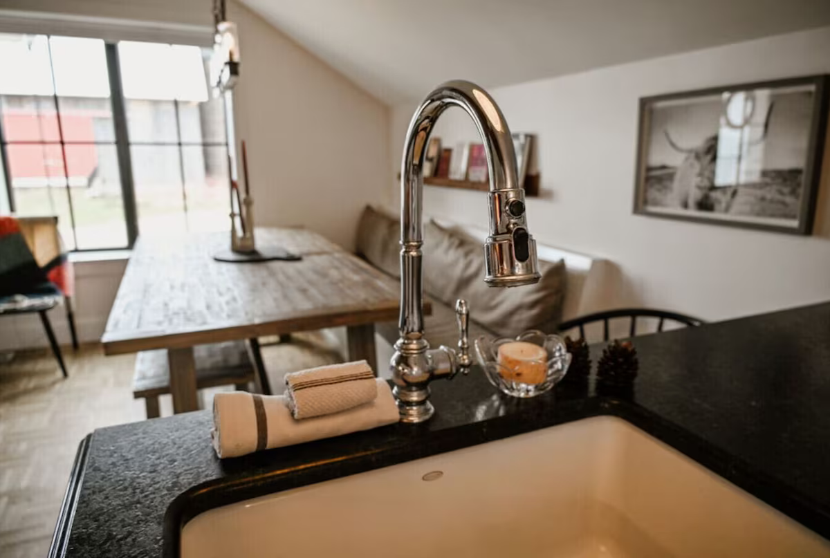Close-up of a kitchen sink with a modern chrome faucet, a candle in a glass bowl, and rolled cloth towels, with a blurred dining area and window in the background.