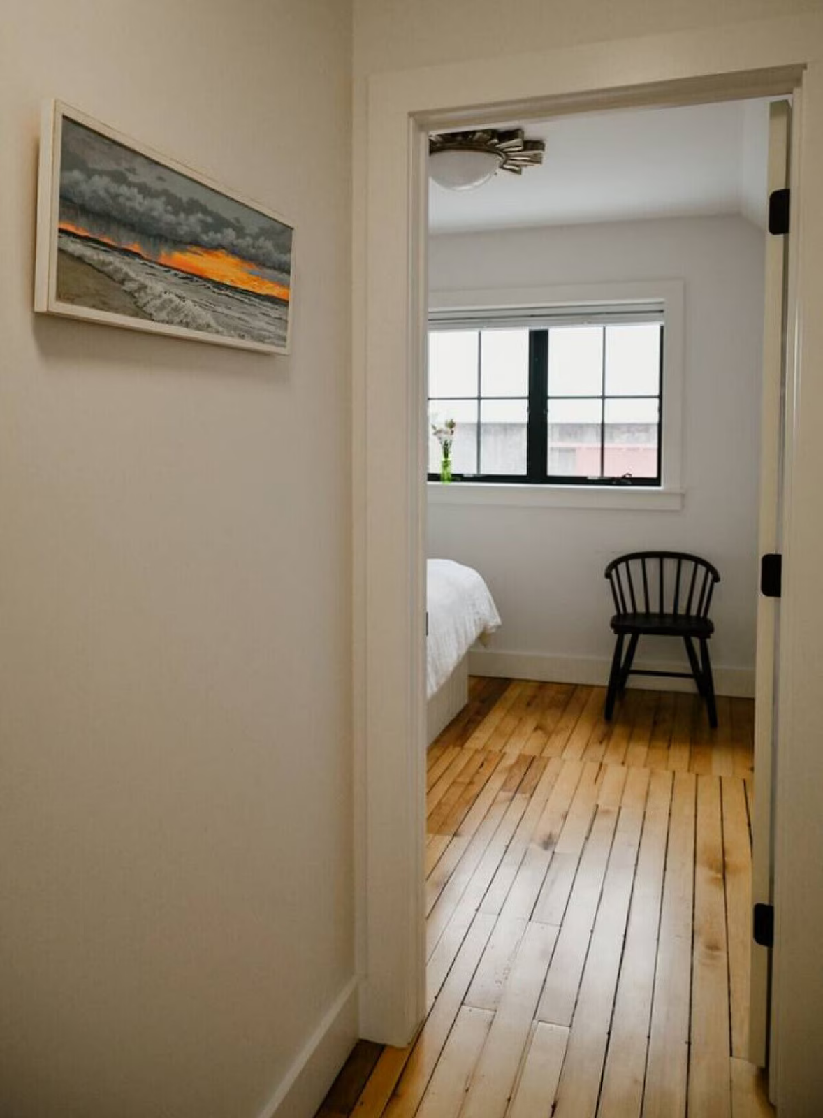 View through a doorway into a bedroom with a wooden floor, a window with blinds, a black chair, and a bed with white bedding, with a sunset landscape painting on the wall.