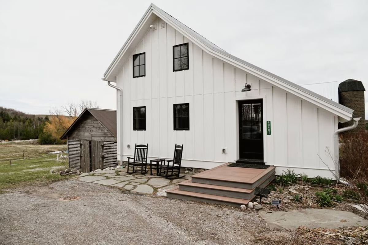 A white two-story house with black window frames and a black front door, with a small set of wooden stairs leading up to the entrance. Two black rocking chairs are positioned outside the house on a stone patio, and a small garden with some plants is 
