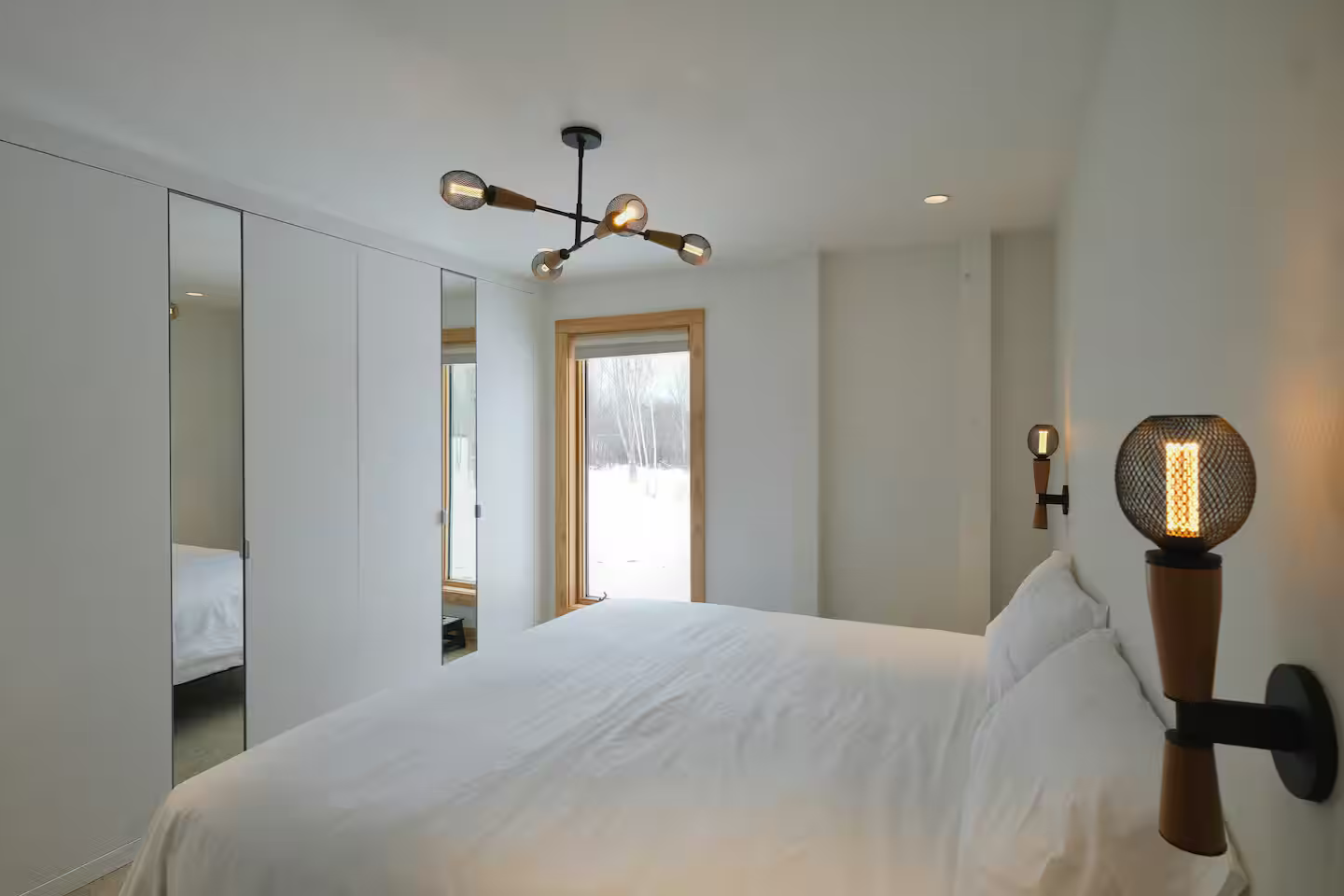 Modern bedroom with white walls, a large bed with white bedding, mirrored closet doors, a window showing a snowy outdoor scene, and modern black and wooden light fixtures.