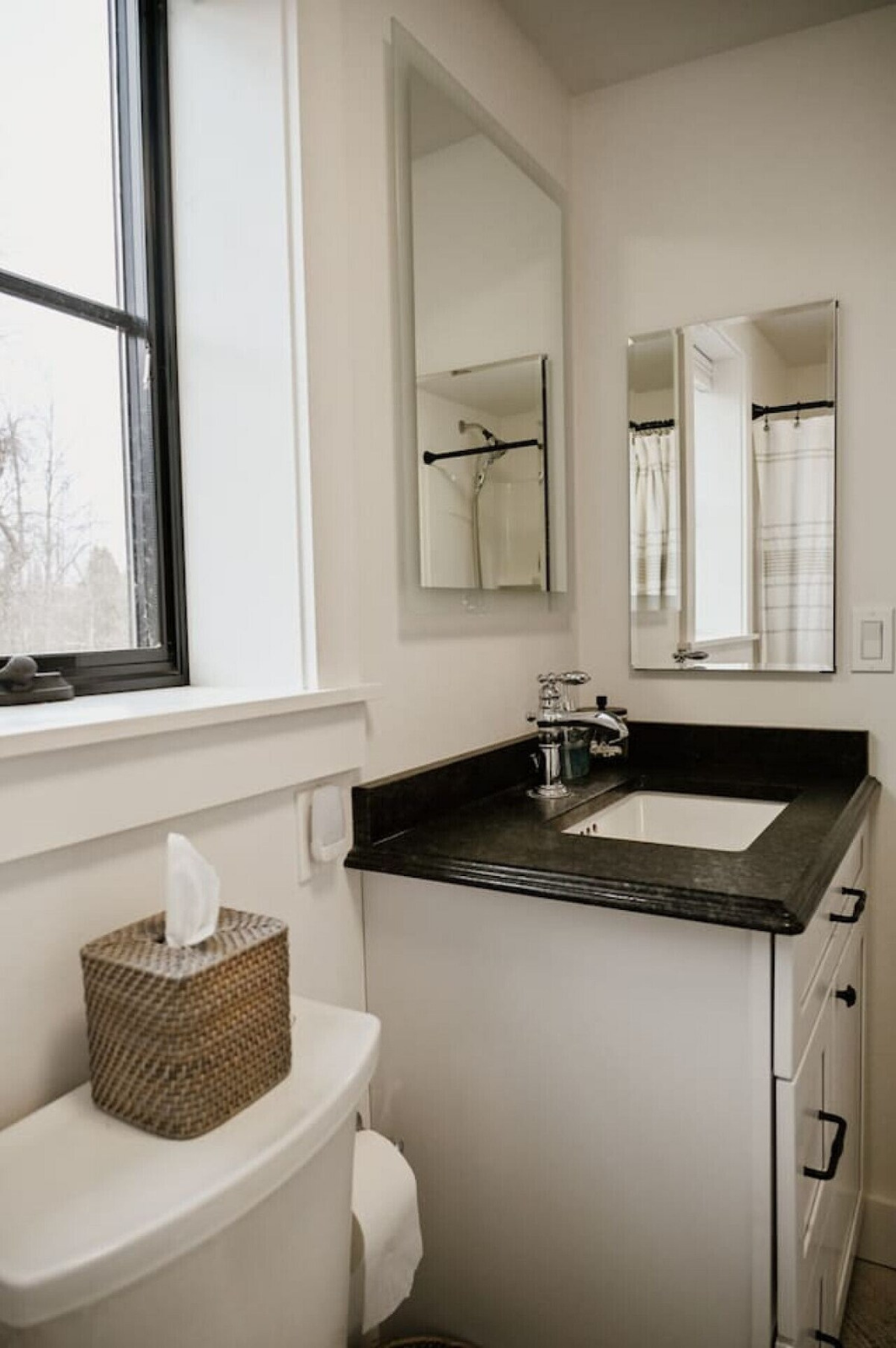 A bathroom with a white toilet, a woven tissue box on top, a black countertop with a sink, a silver faucet, two small mirrors, a window with a view of trees, and a partial shower curtain in the background.