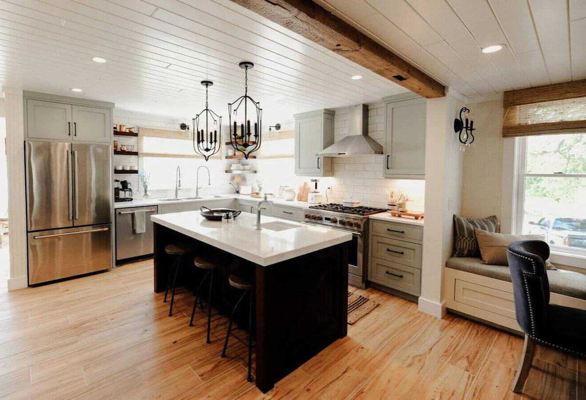 Modern kitchen with stainless steel refrigerator, white countertops, light gray cabinets, gas stove with range hood, island with a sink, pendant lights, wooden floors, and a window seat.