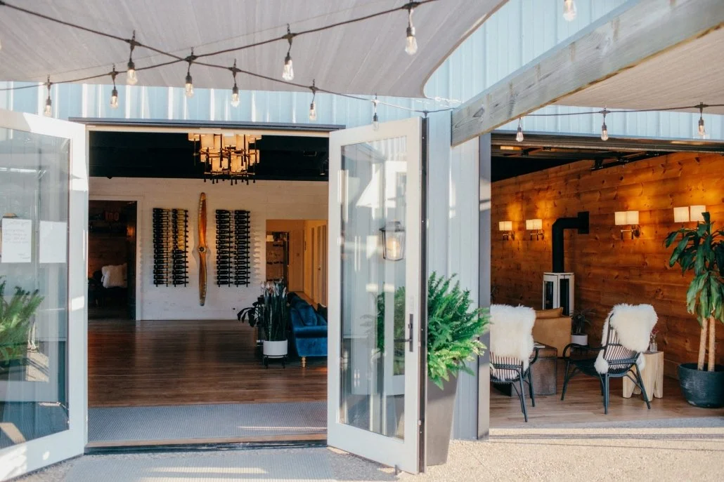 Open doors leading into a modern, well-lit interior space with wooden walls, string lights hanging from the ceiling, and cozy seating with furry throws. The room features warm lighting, indoor plants, and a wine rack on the back wall.