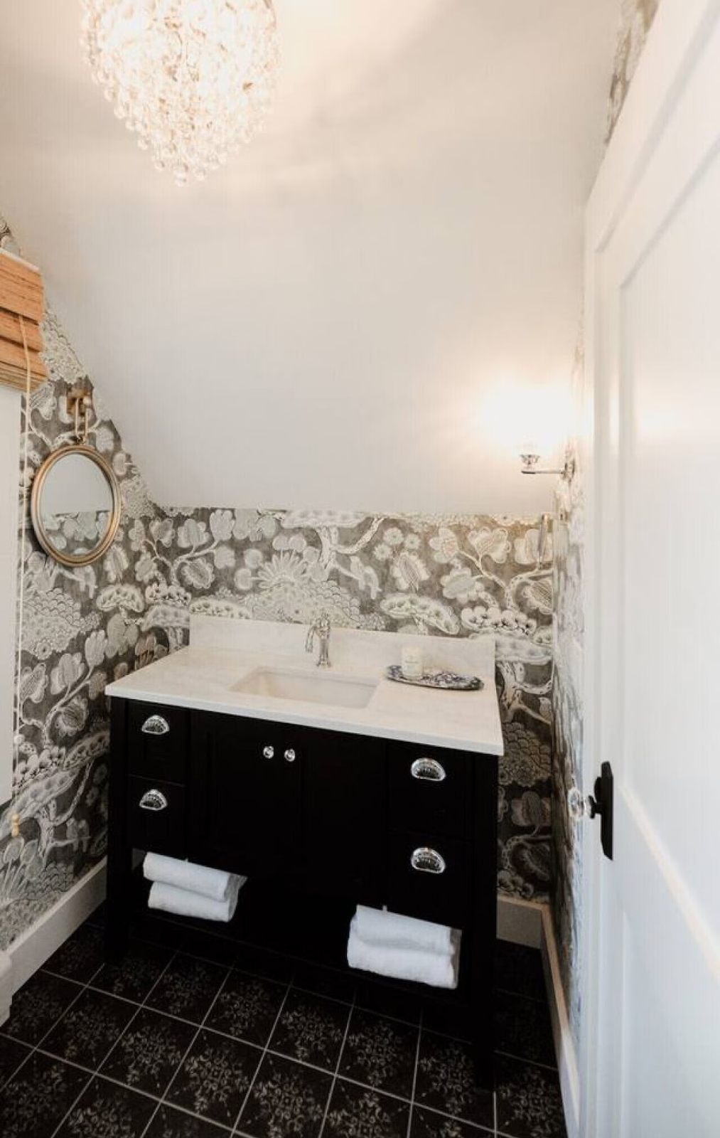 Small bathroom with black vanity, white countertop, and wallpaper with black and white floral pattern. Towels are stored under the sink, and there is a round mirror on the wall. Lighting includes a sconce on each side of the mirror and a ceiling fixt