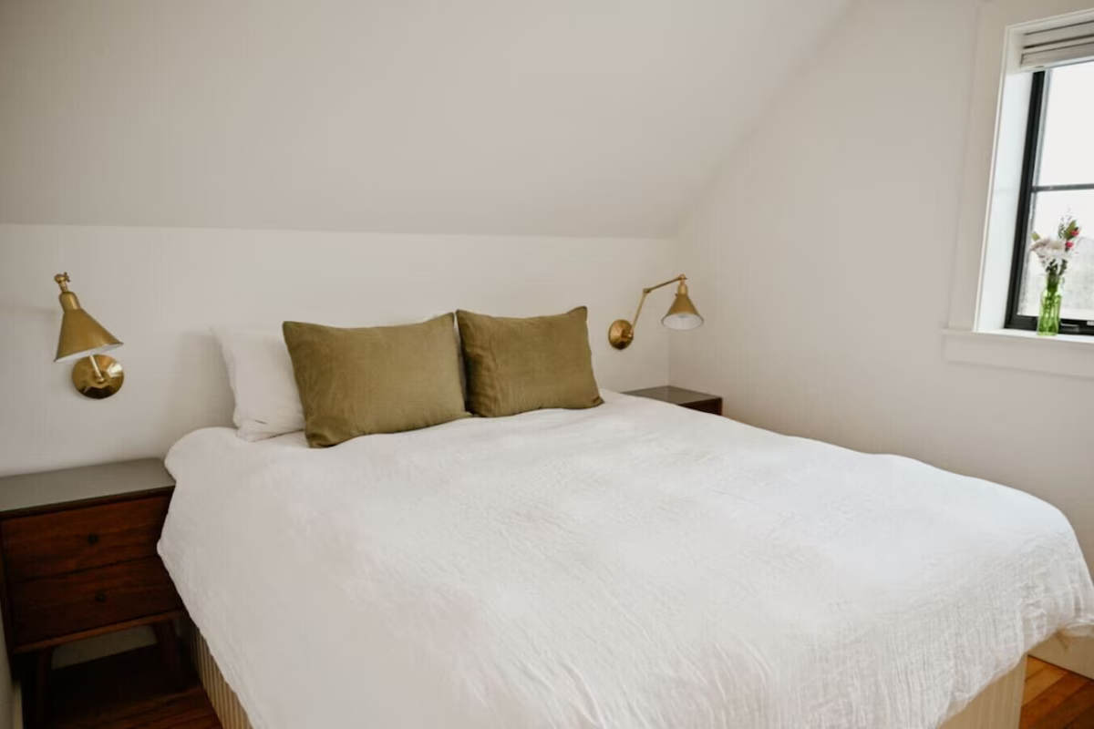 A cozy bedroom with a white bedspread, two white pillows, and three green pillows, with two gold wall-mounted reading lamps, a dark wooden nightstand on each side, and a window with a vase of flowers on the window sill.
