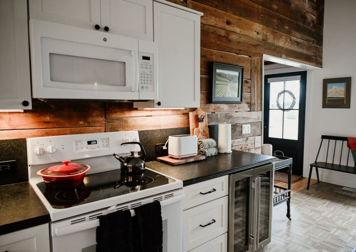 Kitchen with white cabinets, black countertop, and wood-paneled wall. Includes a microwave, stove with a red pot, toaster, paper towels, and a small refrigerator. There is a door with glass panel and a wreath outside, and a small bench and picture on
