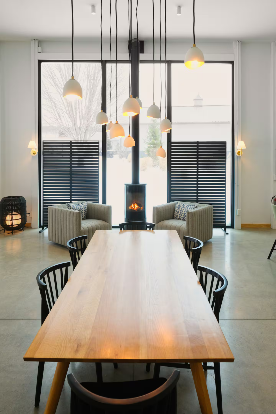Modern interior with a long wooden dining table, black chairs, a fireplace, and hanging pendant lights, large windows with black slatted screens, and armchairs with cushions in the background.