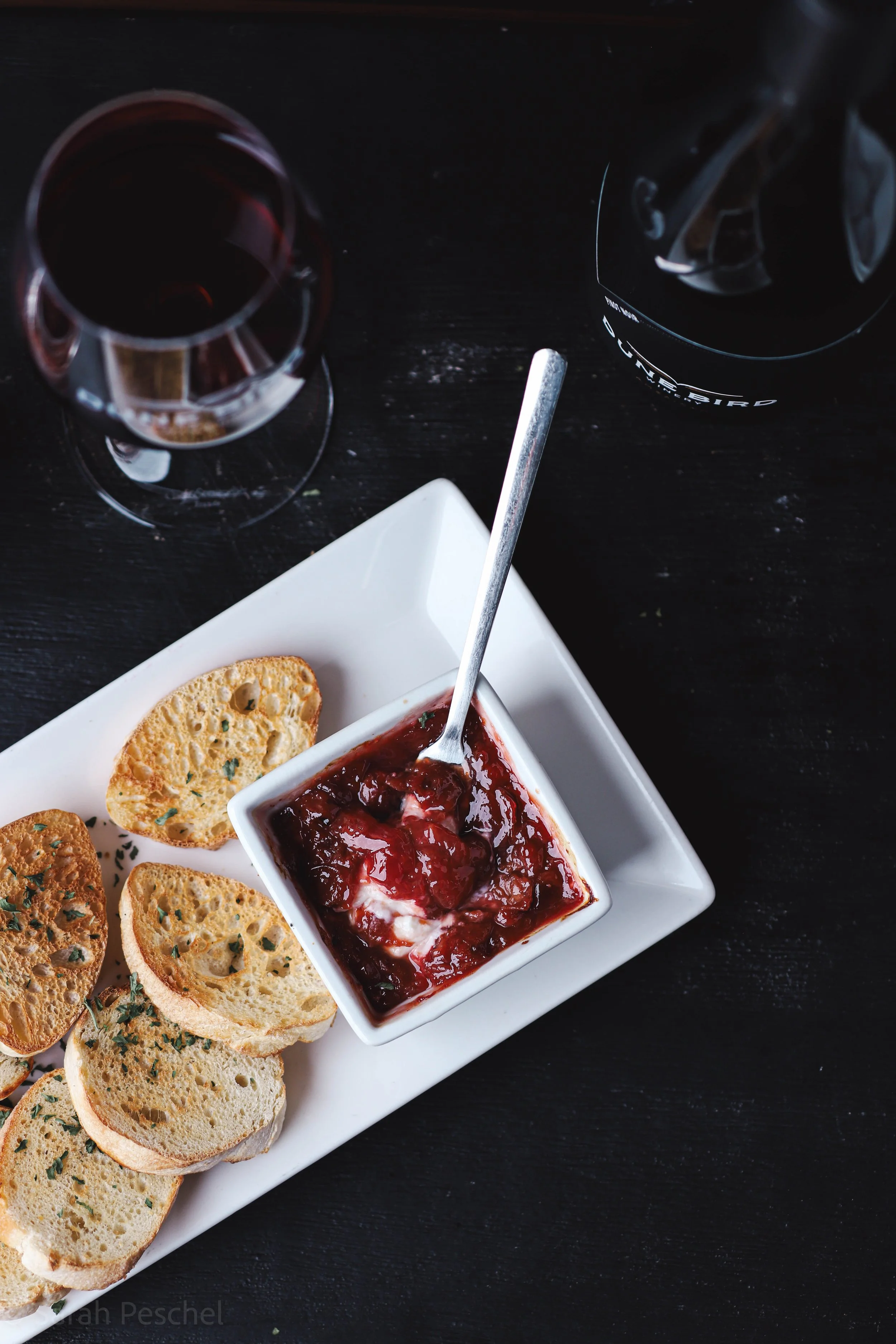 A plate of toasted baguette slices garnished with herbs, a small white bowl filled with red jam or jelly with a spoon, a glass of red wine, and a bottle of wine on a dark surface.