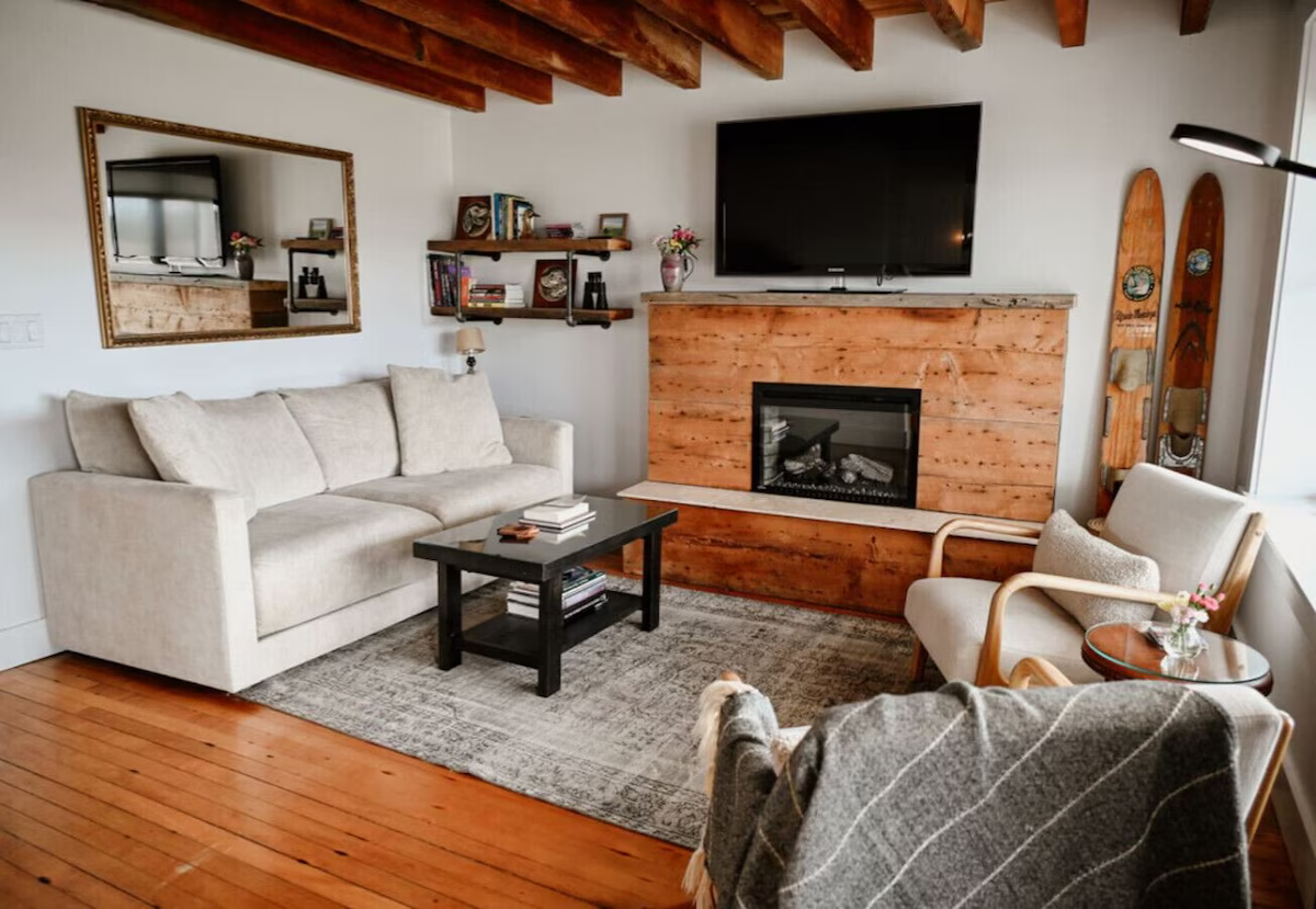 Living room with a beige sofa, a black coffee table, a rug, a wooden fireplace with a TV mounted above, a armchair with a throw blanket, and a small round side table with flowers. There are wooden ceiling beams, a large mirror, a wall-mounted bookshe