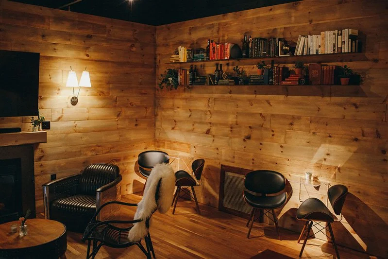 Cozy living room with wooden walls, a wall-mounted lamp, a bookshelf, and several black armchairs and chairs arranged around the room.
