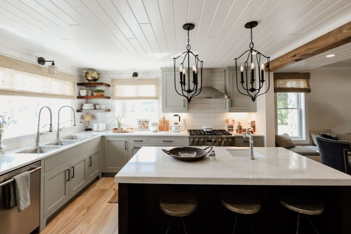 Modern kitchen with gray cabinetry, white countertops, and a white tile backsplash, featuring a central island with stools and decorative chandeliers hanging from the ceiling.