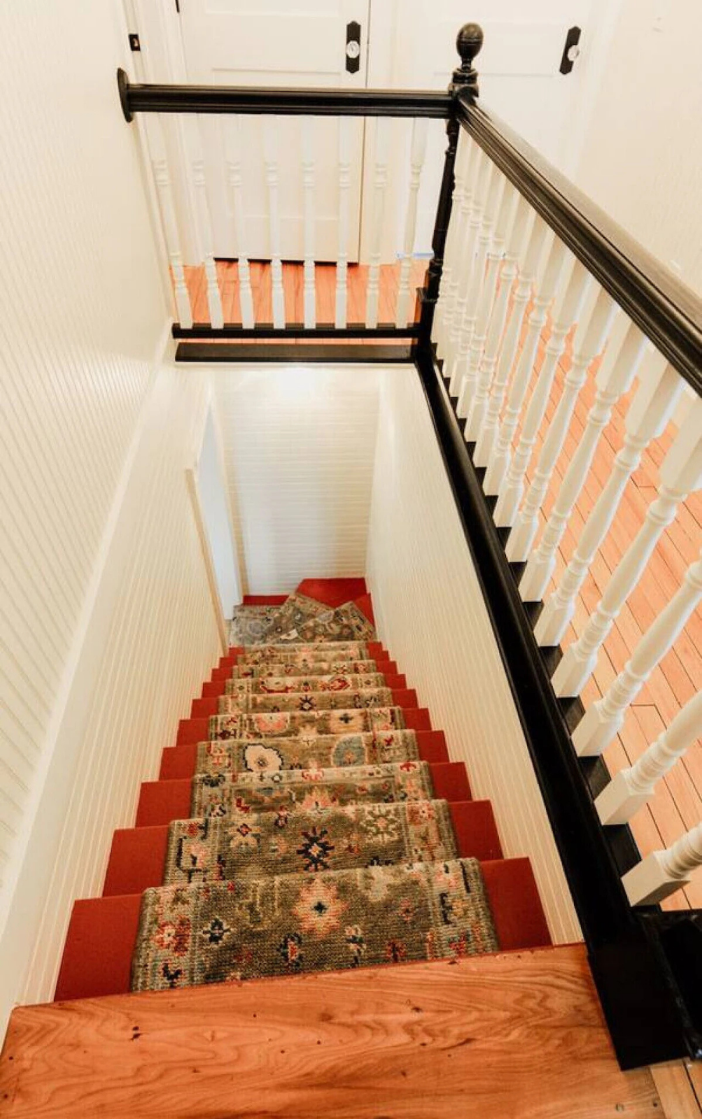View of a staircase with red carpeted steps, a floral patterned rug runner, white walls, and a wooden and white railing, looking downward from the top.