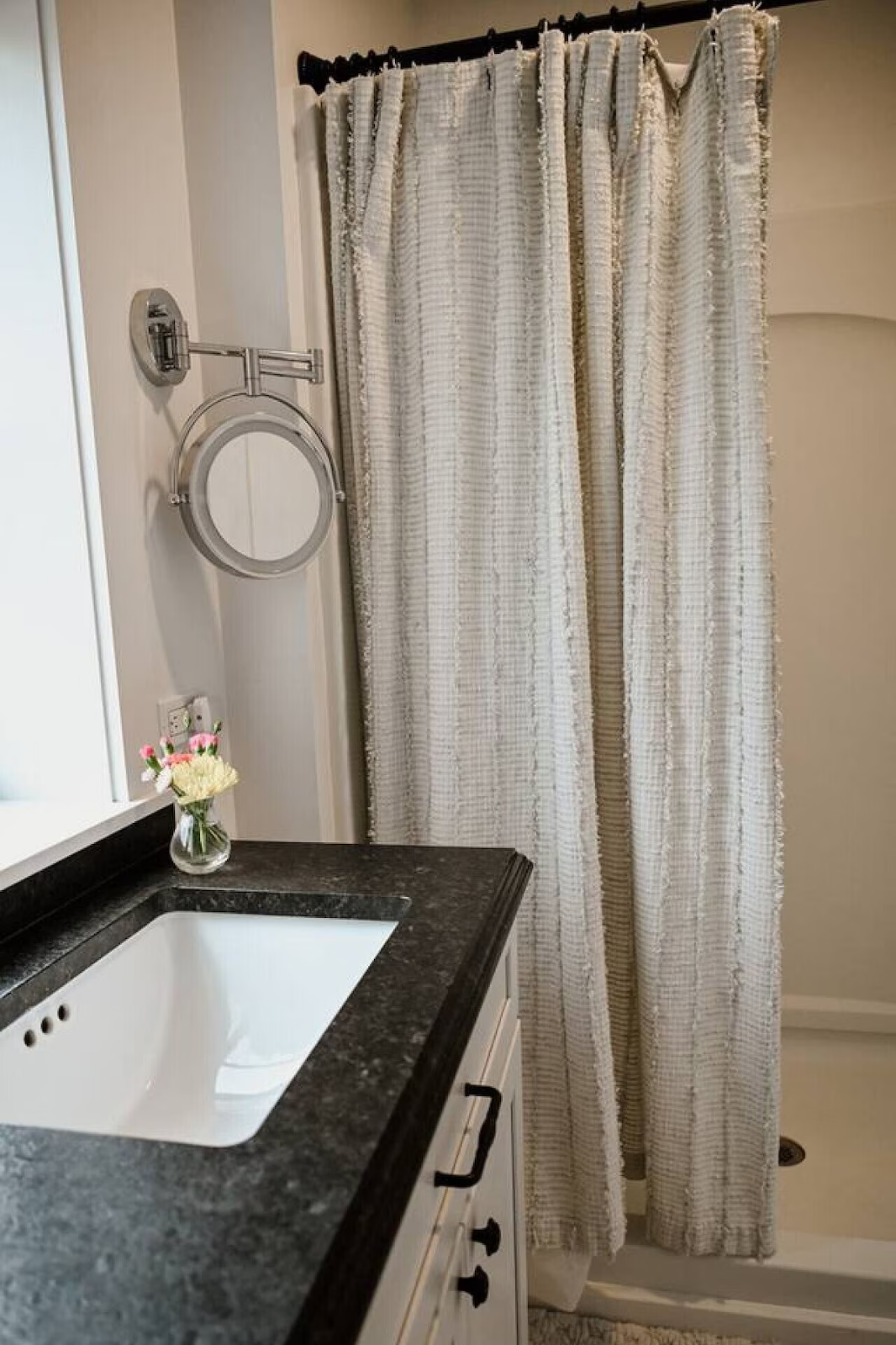 Bathroom with a white and black granite countertop, a white sink, a small vase with pink and white flowers, a wall-mounted mirror with a silver frame, and a white shower curtain with textured stripes.