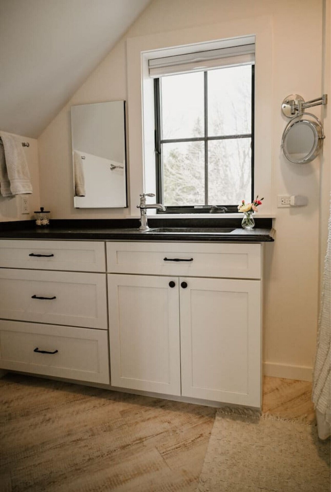 A bathroom vanity with white cabinets, black handles, black countertop, a silver faucet, a window with white frame, a small mirror, and a small vase with flowers.