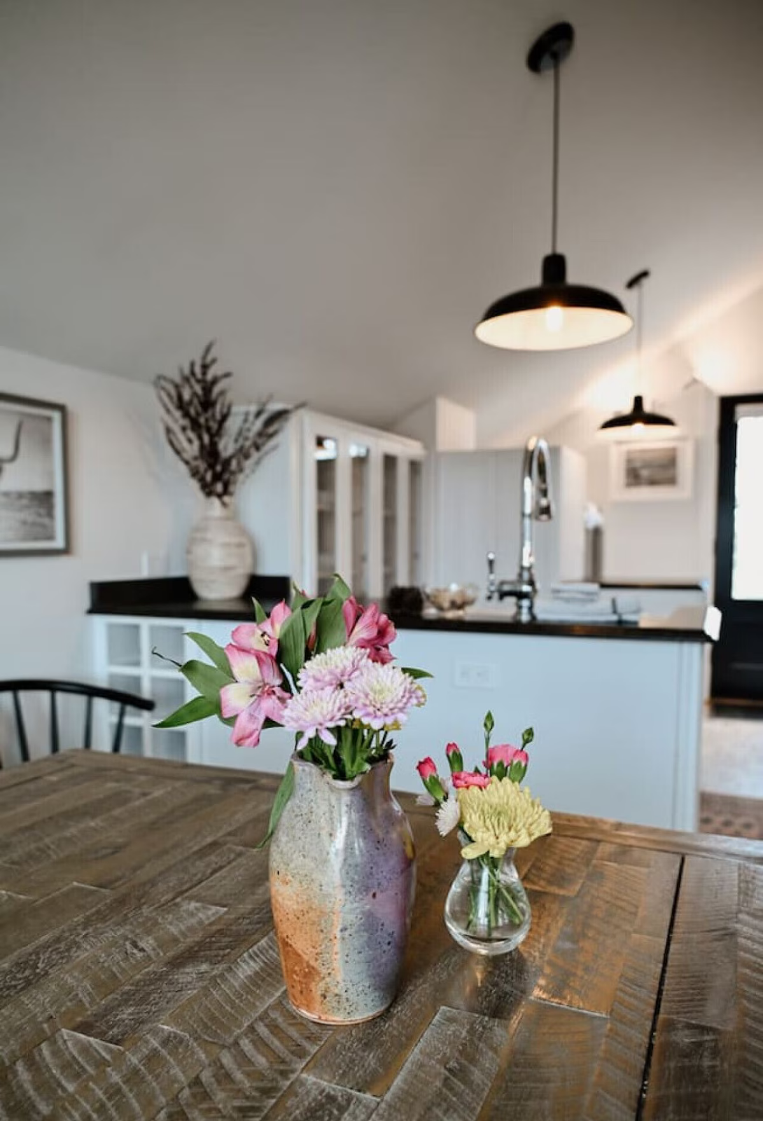 Close-up of a wooden dining table with two vases of flowers, one with pink and white flowers and the other with yellow and pink flowers, in a well-lit kitchen and dining area.