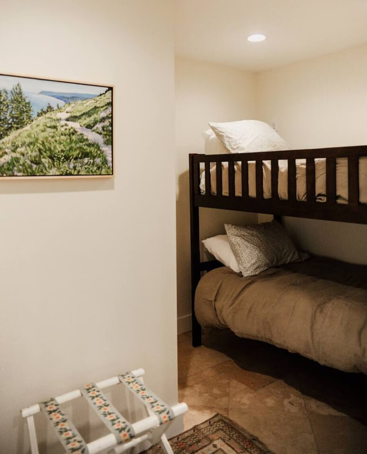 A small room with beige walls, featuring a bunk bed with dark wooden frame. The top bunk has a white pillow and beige bedding, the bottom bunk has a gray pillow and similar bedding. To the left, there is a framed landscape painting of a mountain path