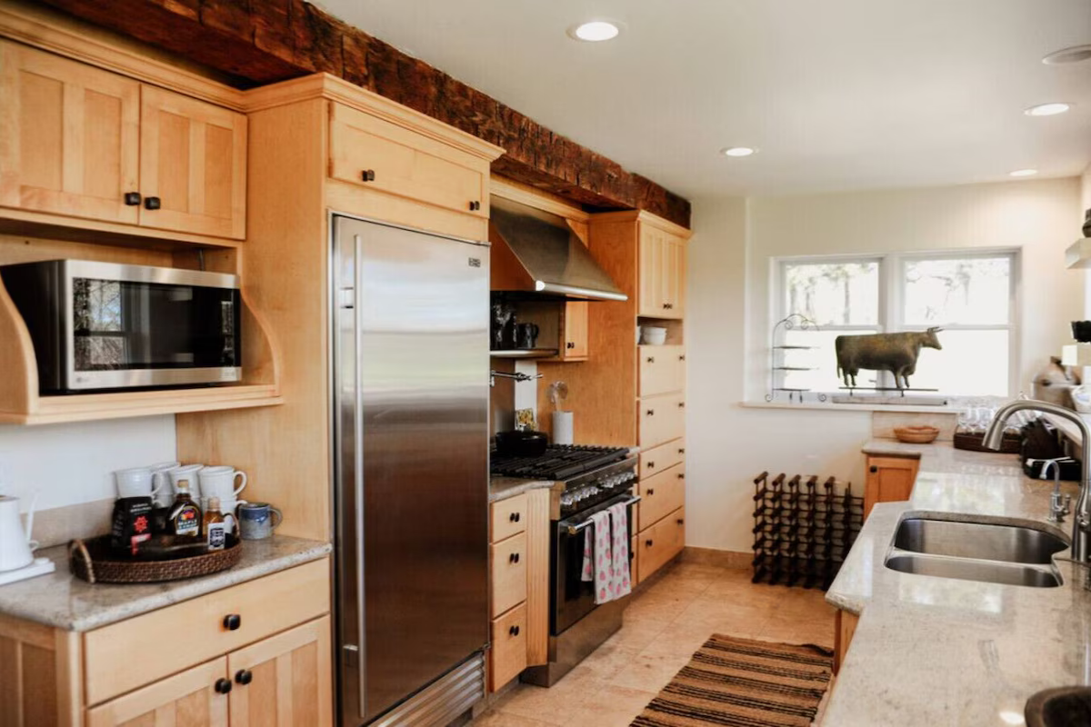 Kitchen with light wood cabinets, stainless steel refrigerator, microwave, stove, beige countertops, and a window with a decorative cow sculpture on the sill.