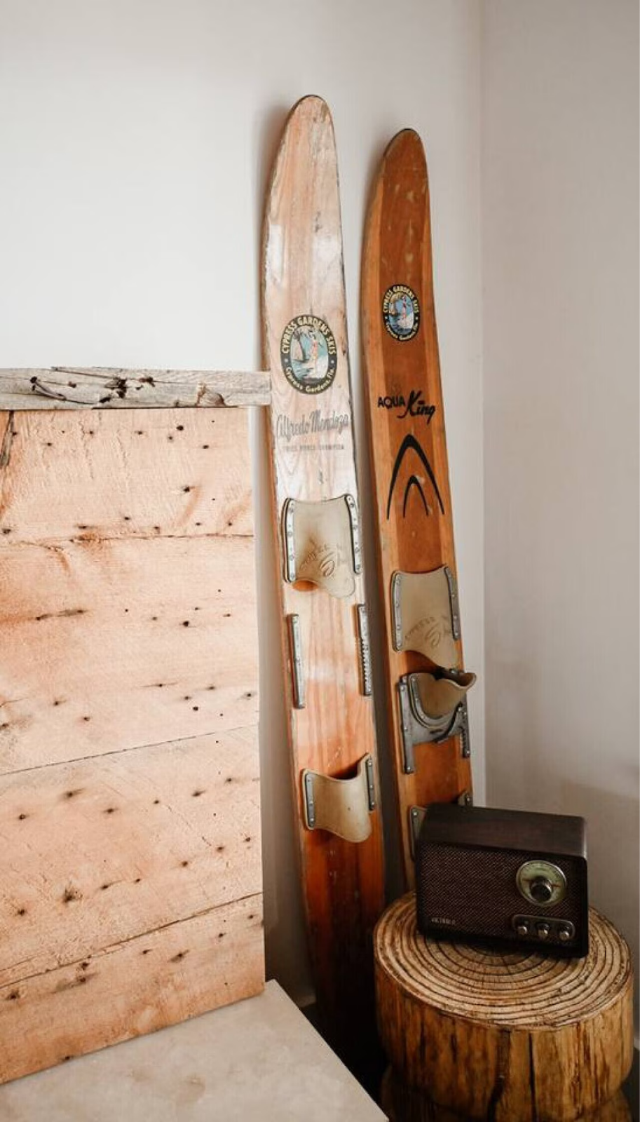 Two vintage wooden skis with bindings, leaning against a wall beside a wooden stump with a vintage radio on top.