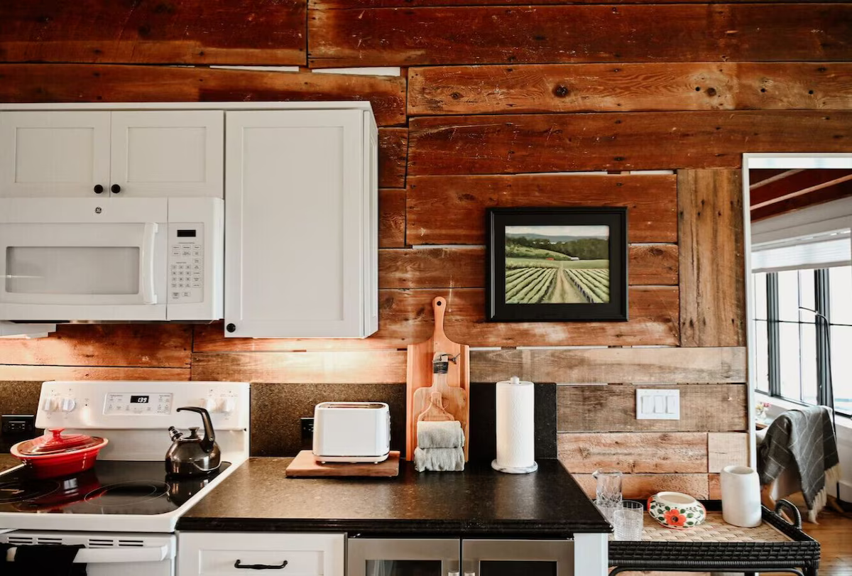 Kitchen with white cabinets, black countertop, wooden wall, and a framed landscape picture of a farm field.