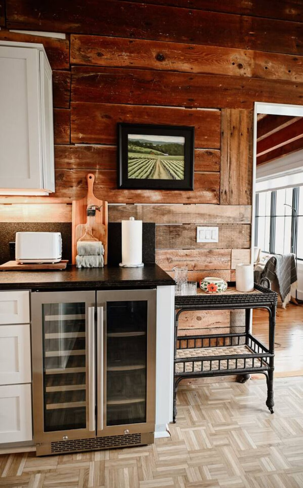 Interior view of a kitchen with a rustic wooden wall, a black countertop, a small wine cooler with glass doors, a toaster, paper towels, a framed landscape painting, and a small black table with a decorative bowl.