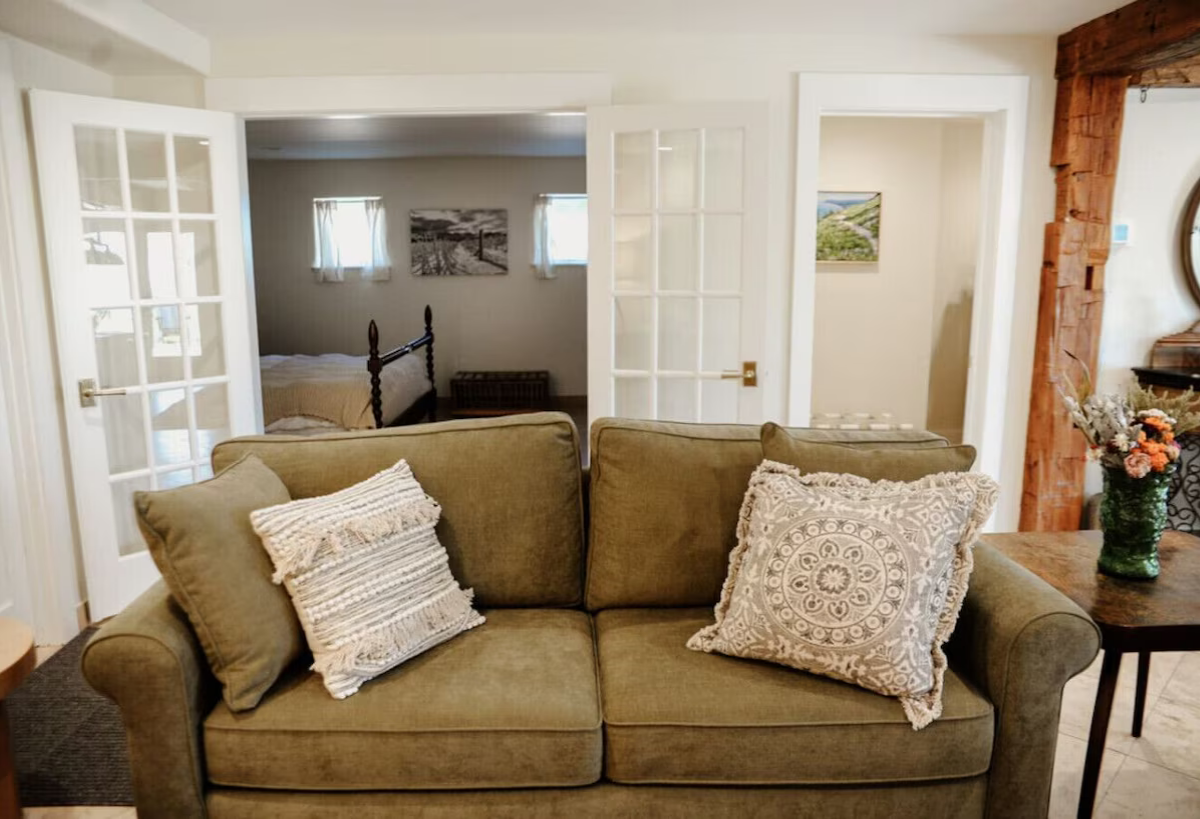 Living room with a green couch and patterned pillows, leading to a bedroom with a wooden bed frame and wall art, with glass double doors and a partial view of a dining area with a floral arrangement.