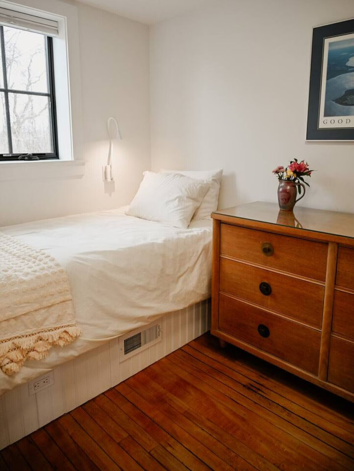 A cozy bedroom with a single bed covered in white bedding, next to a wooden dresser with a vase of flowers, a framed picture on the wall, and a window with a view of leafless trees outside.