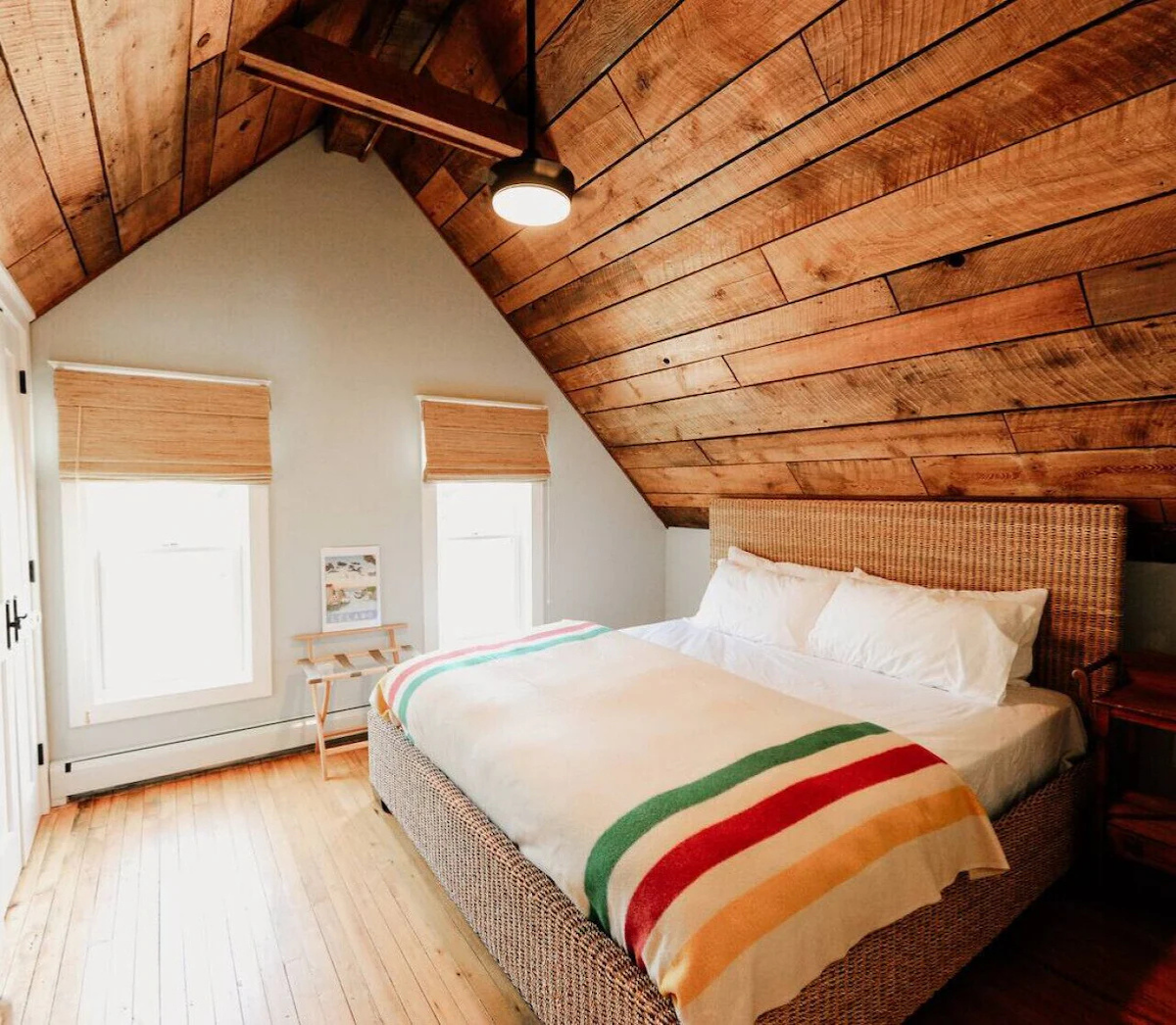 A cozy bedroom with a sloped wooden ceiling, a bed with white linens and a colorful striped blanket, two small windows with blinds, and a wooden nightstand.