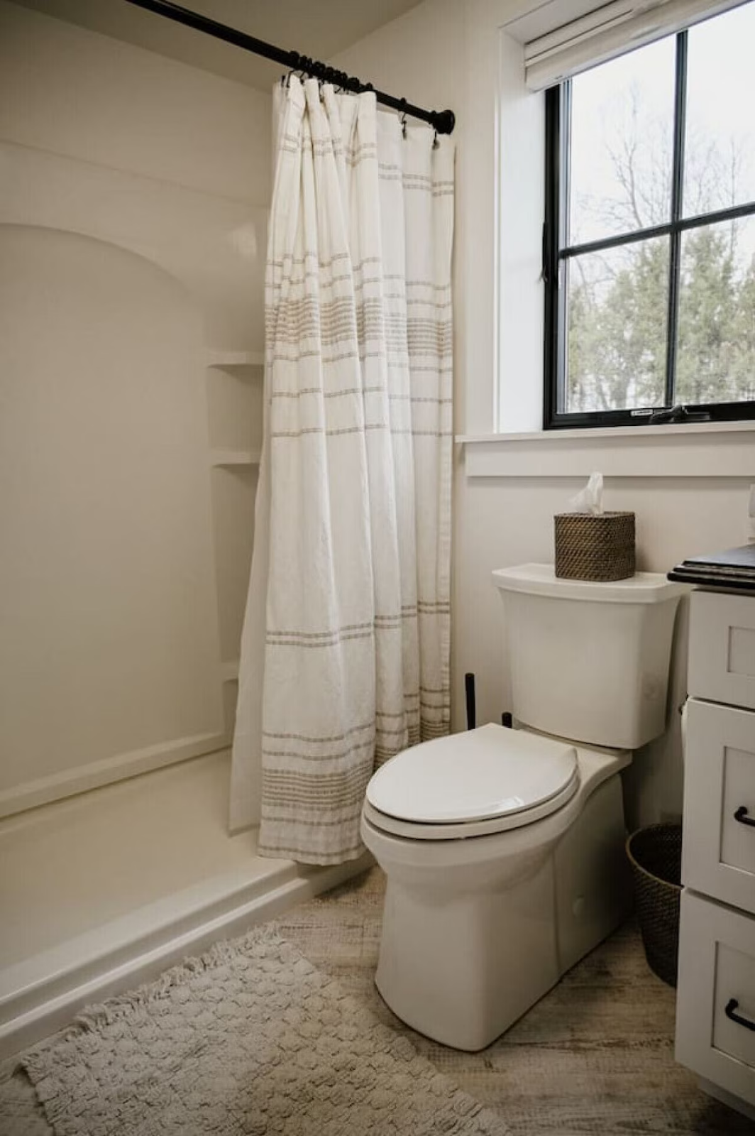 A bathroom with a shower, white curtain, toilet, window, and a small brown basket.