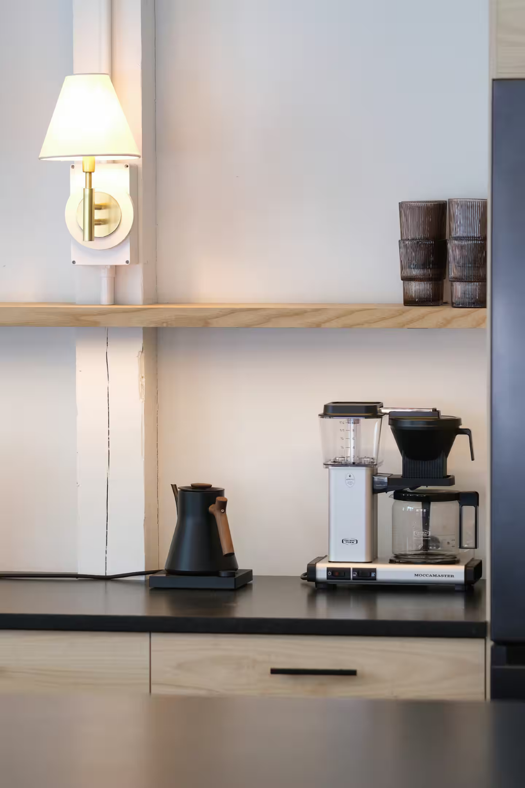 Kitchen countertop with a black electric kettle, a coffee drip machine, a blender, and a coffee pot, with a wall-mounted lamp and a shelf holding glass cups in the background.