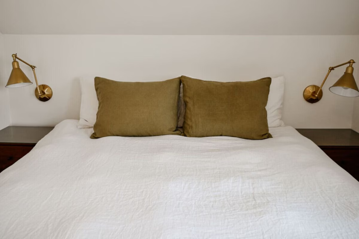 A neatly made bed with white bedding, two large brown pillows, and two white pillows, flanked by two gold-colored wall-mounted reading lamps in a bedroom with a neutral color scheme.