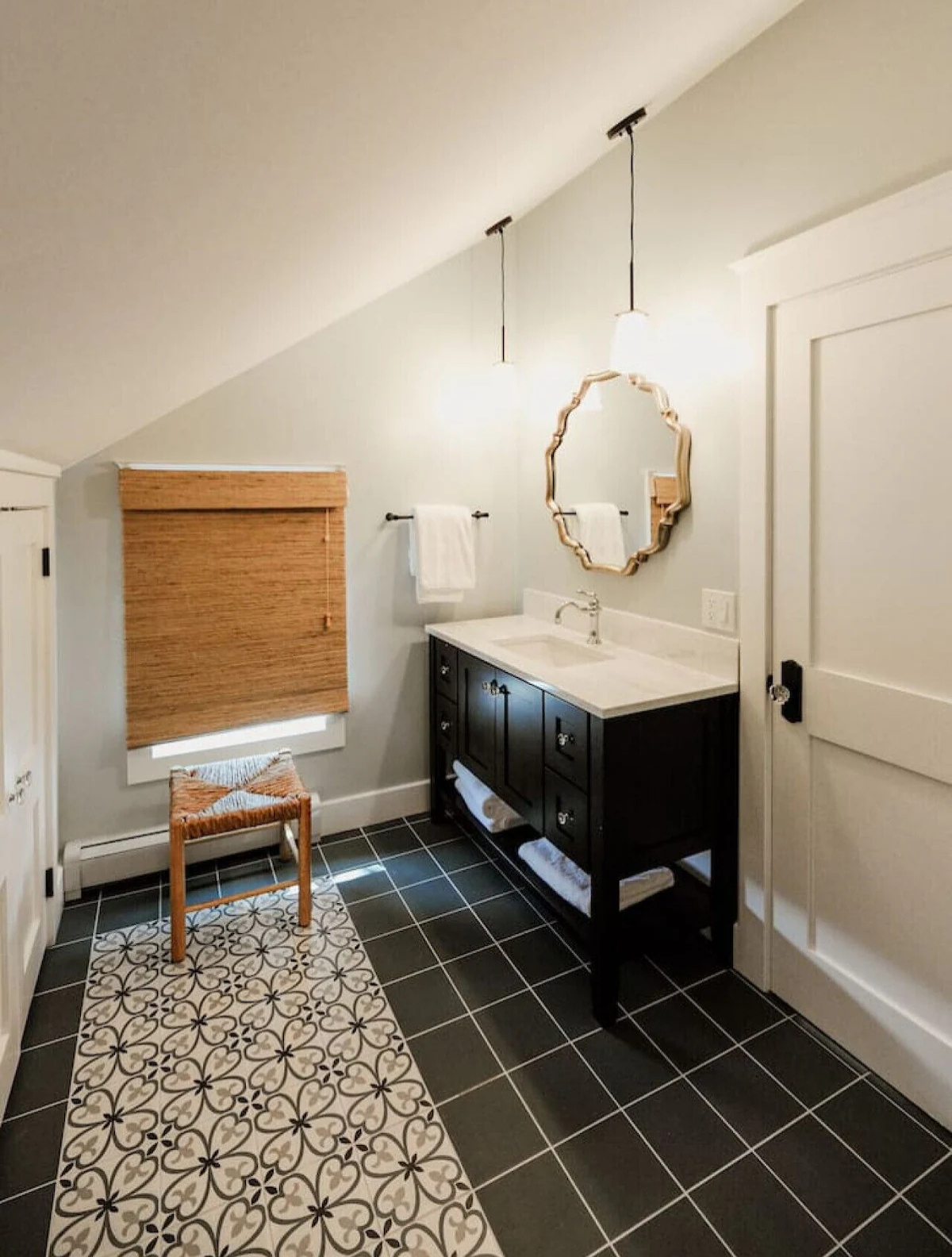 Bathroom with black vanity, white countertop, round mirror, light fixtures, towel rack, window with beige blinds, decorative rug, white door, sloped ceiling, and wood accents.