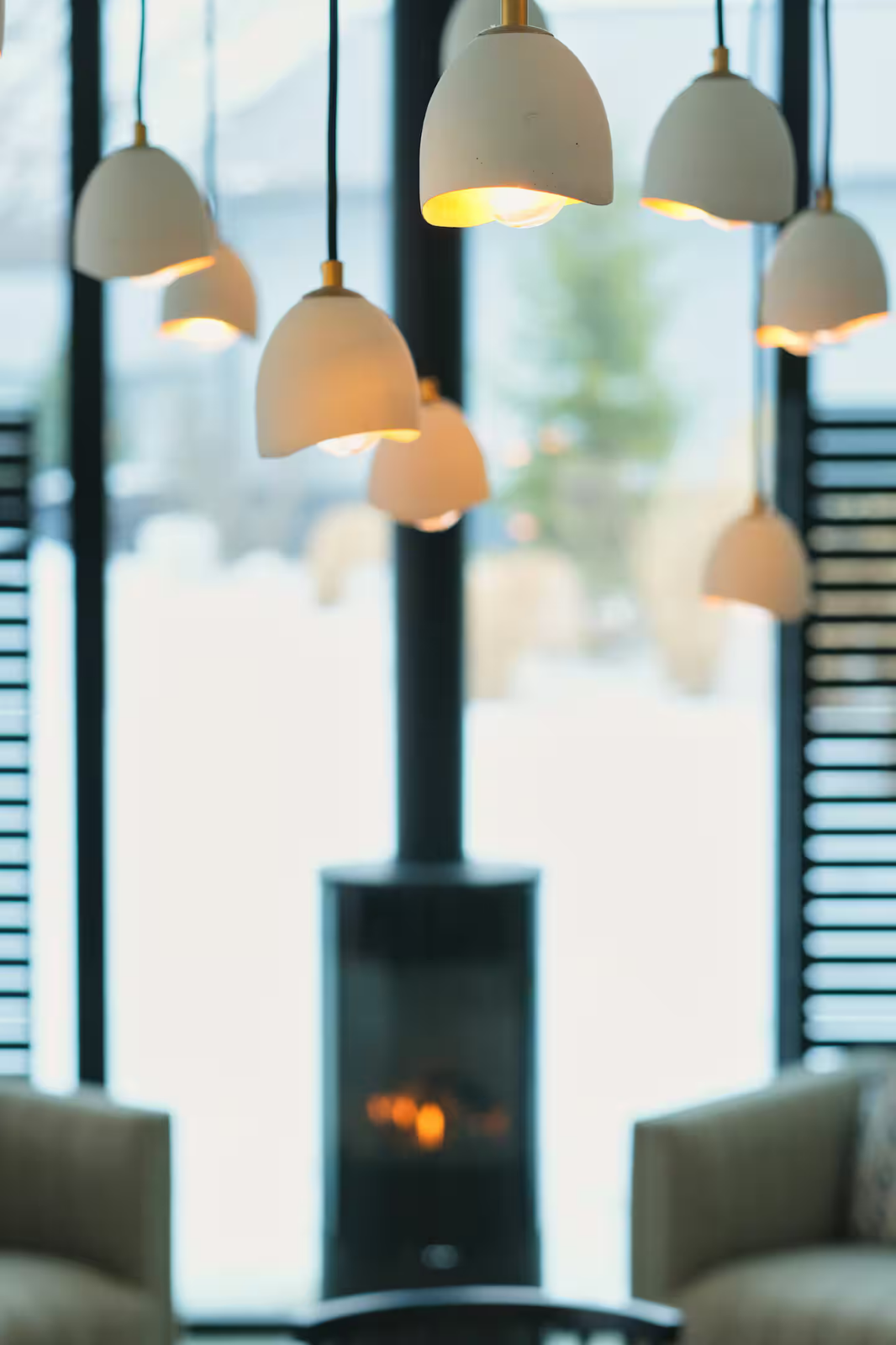 Indoor space featuring multiple hanging pendant lights with warm glow, large window or glass door with black frames, and a dark fireplace or stove with visible glowing embers.