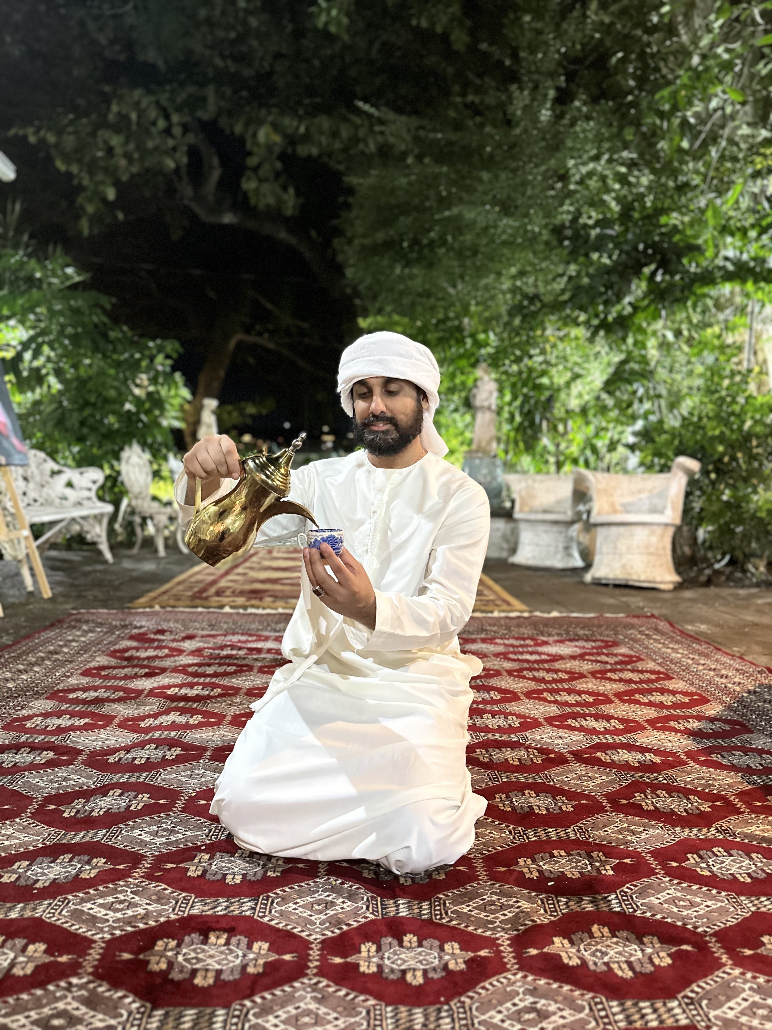 A man dressed in traditional white attire and head covering is sitting on a decorative red patterned rug outdoors, pouring tea from a golden teapot into a small blue and white cup amidst lush green trees and outdoor furniture.