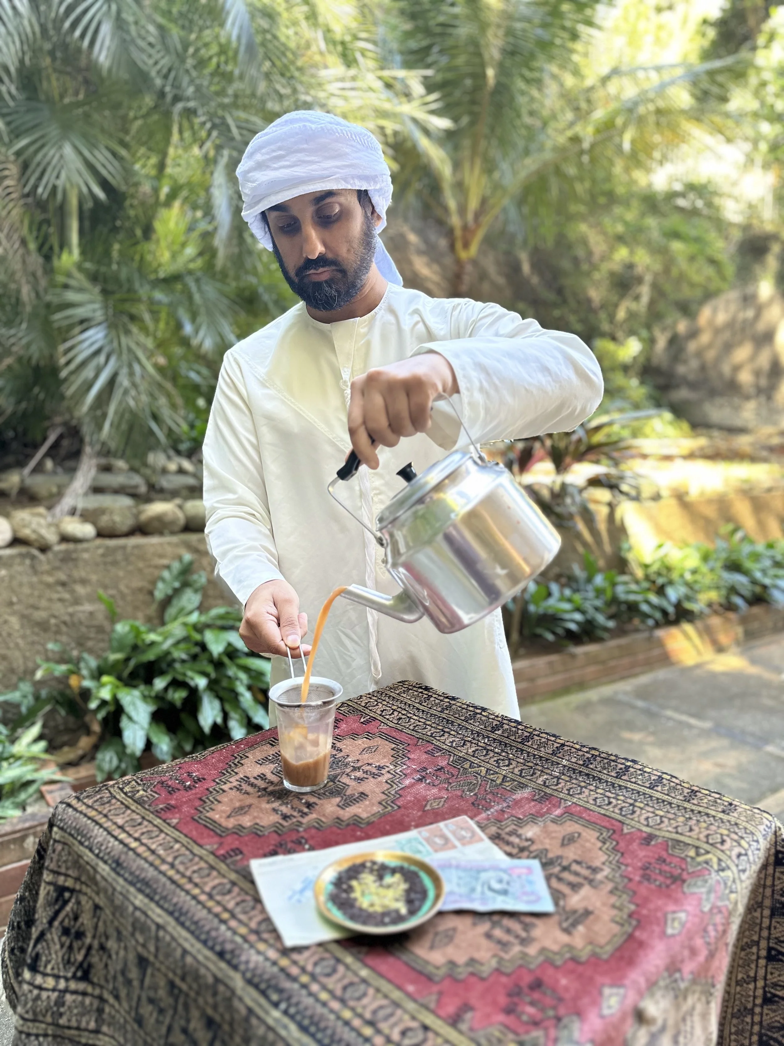 A man dressed in traditional Middle Eastern attire is pouring iced coffee from a metal pot into a glass on a patterned table outdoors, with lush green plants in the background.