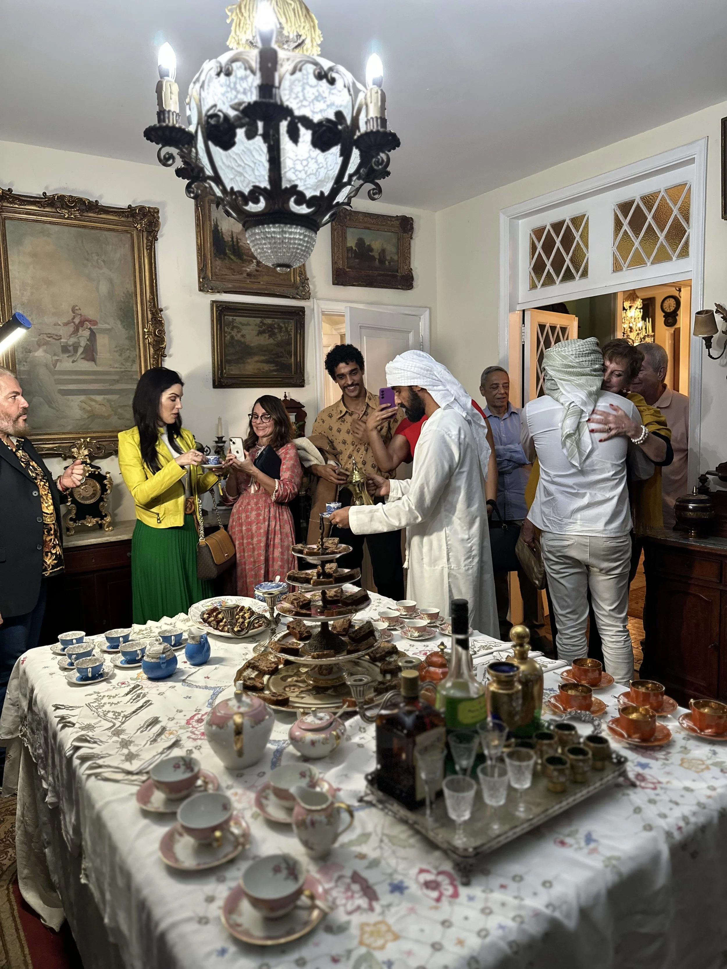 A gathering of diverse people around a table with tea sets, snacks, and beverages in a decorated room with paintings and a chandelier.