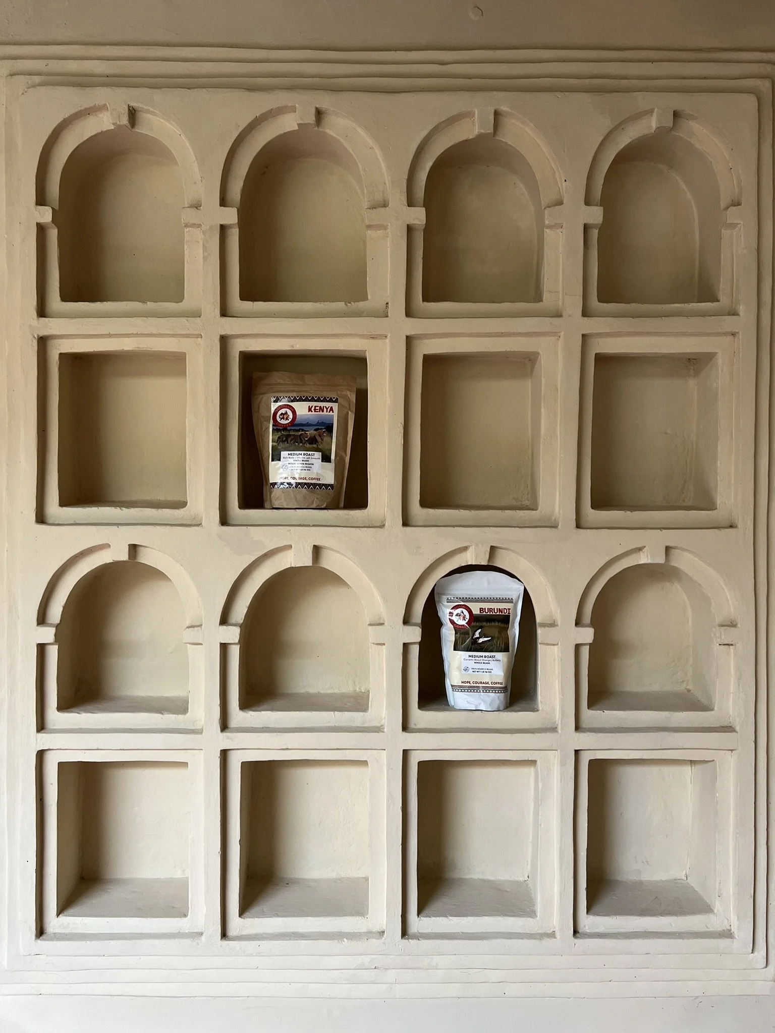 A beige decorative wall with multiple arched and square niches, two of which contain bags of coffee labeled 'Kenya' and 'Burundi'.