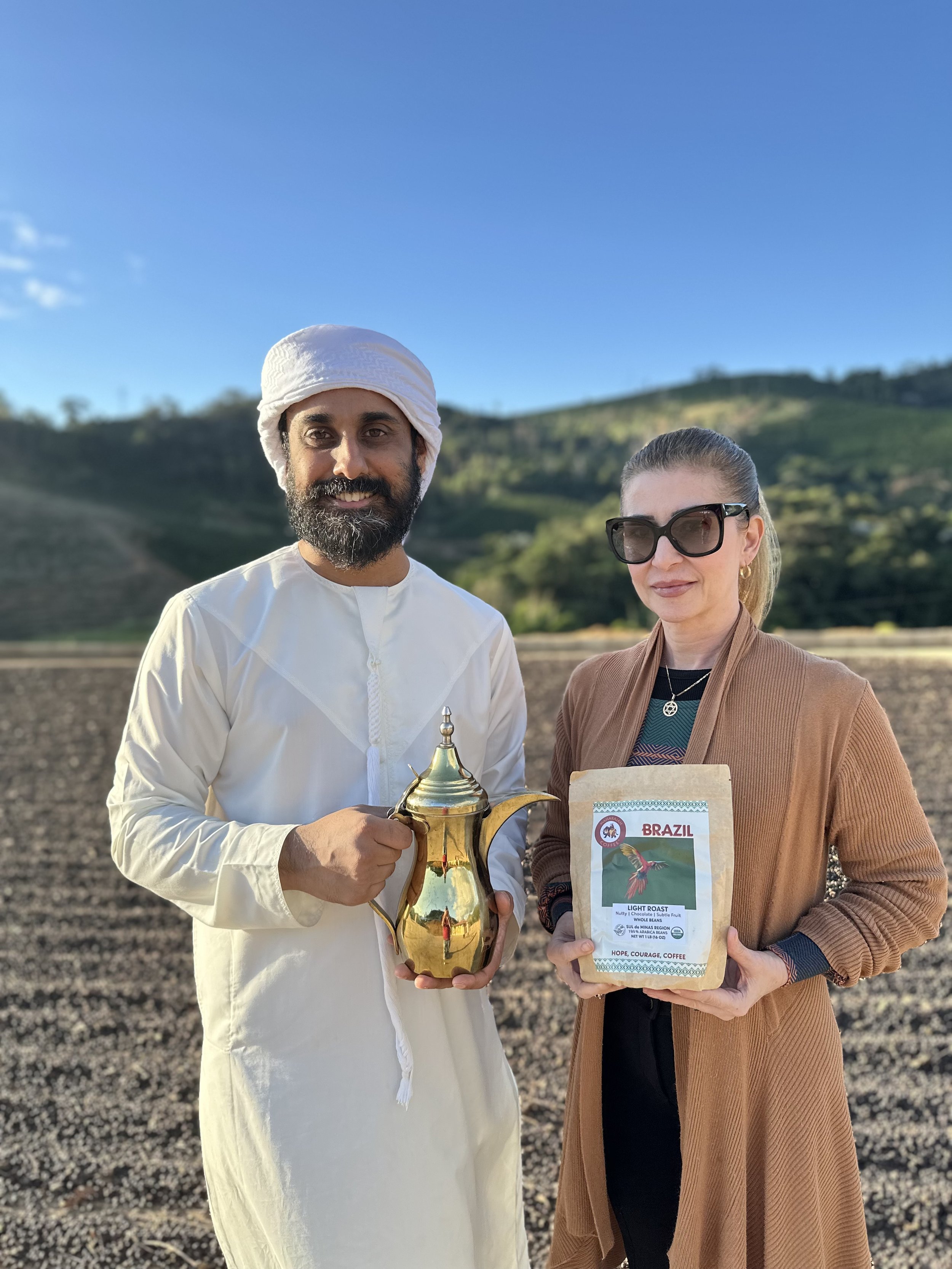 Two people, a man in traditional white Middle Eastern attire and a woman wearing sunglasses and a brown coat, standing outdoors in a field with hills in the background. The man is holding a decorative gold coffee pot, and the woman is holding a bag of Brazilian coffee beans.