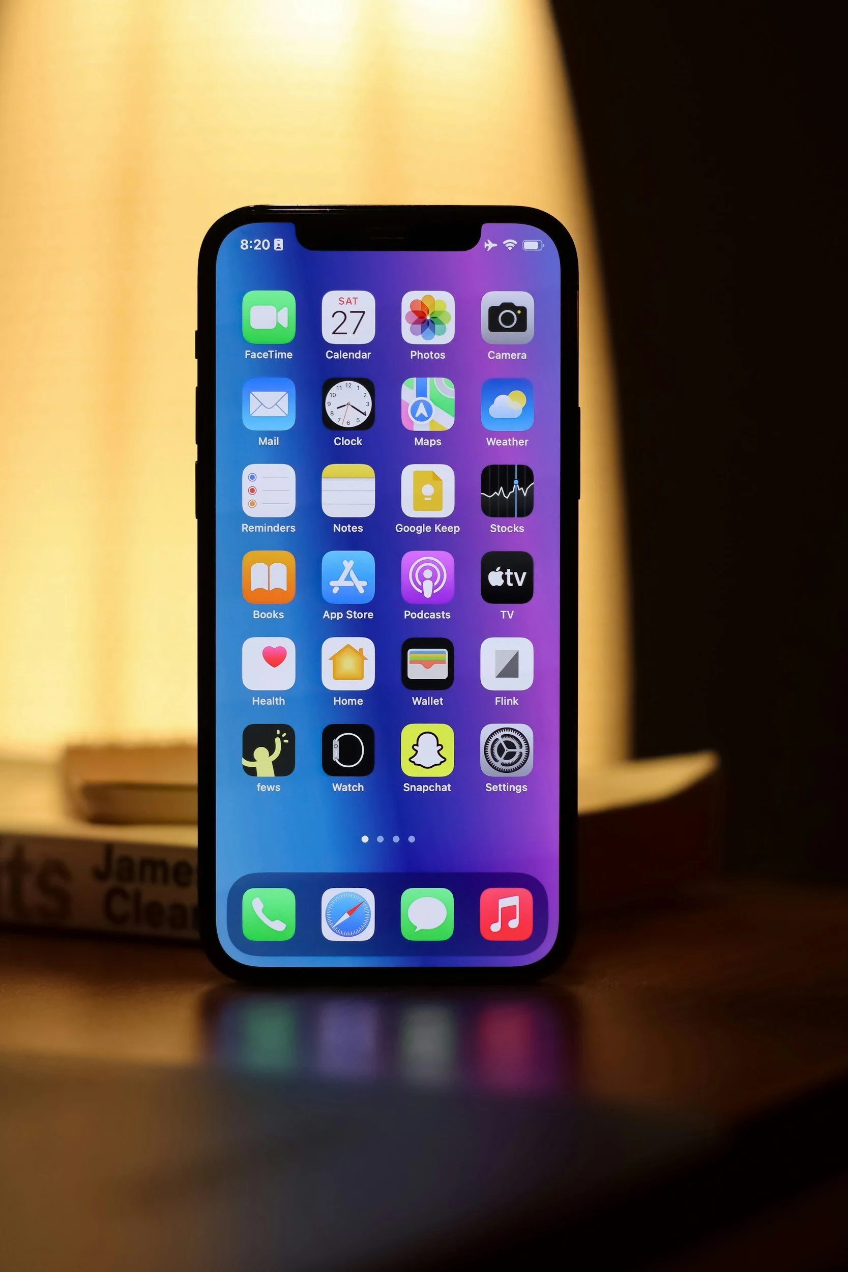 An iPhone with the home screen displaying various app icons, standing on a surface with a blurred background.