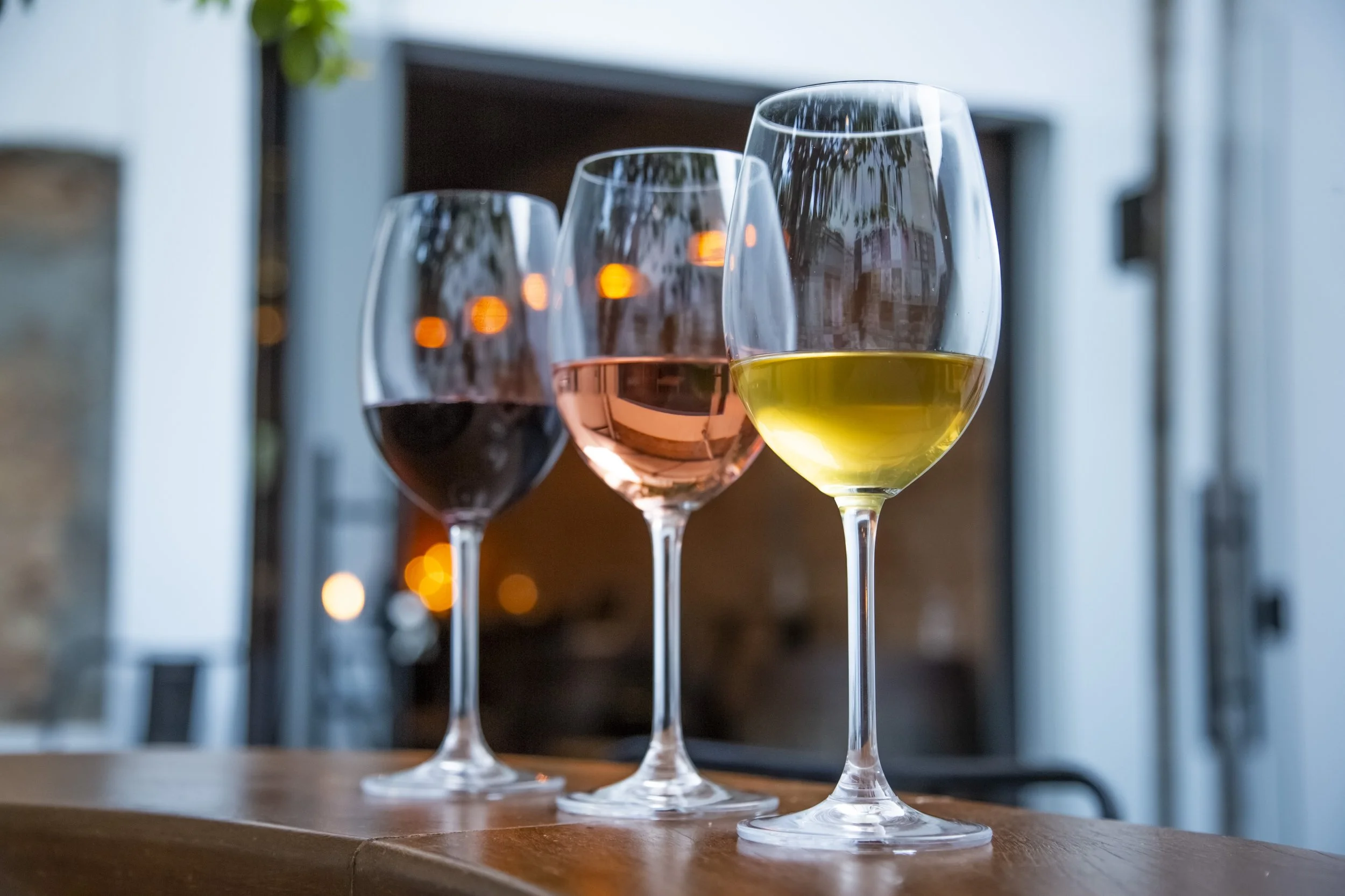 Três taças de vinho ao lado de uma mesa de madeira, com fundo de ambiente interno com iluminação suave e decoração moderna