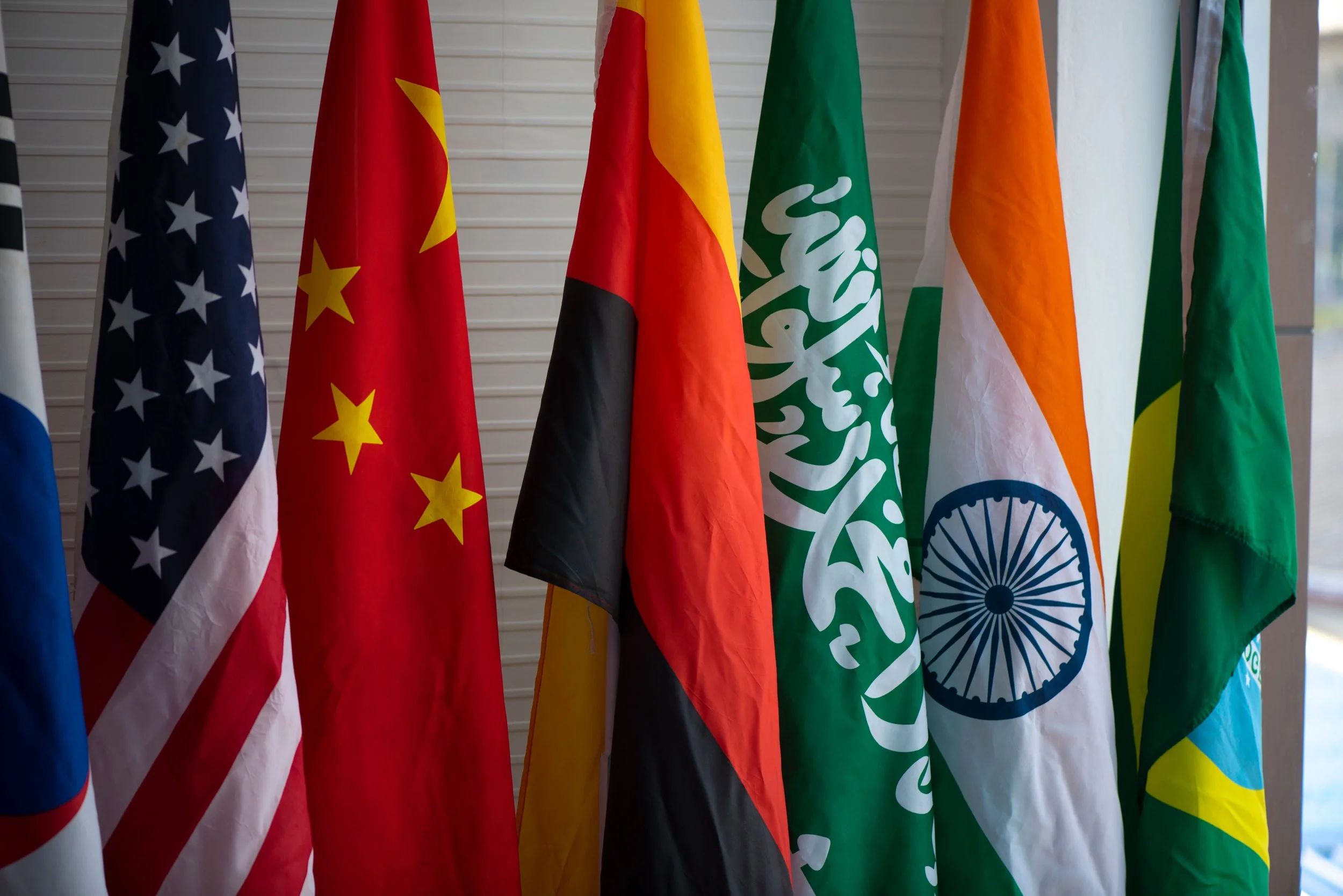 A line of flags - South Korea, U.S., China, Saudi Arabia, India, Brazil - stand next to each other in front of a wall. International Missions Support