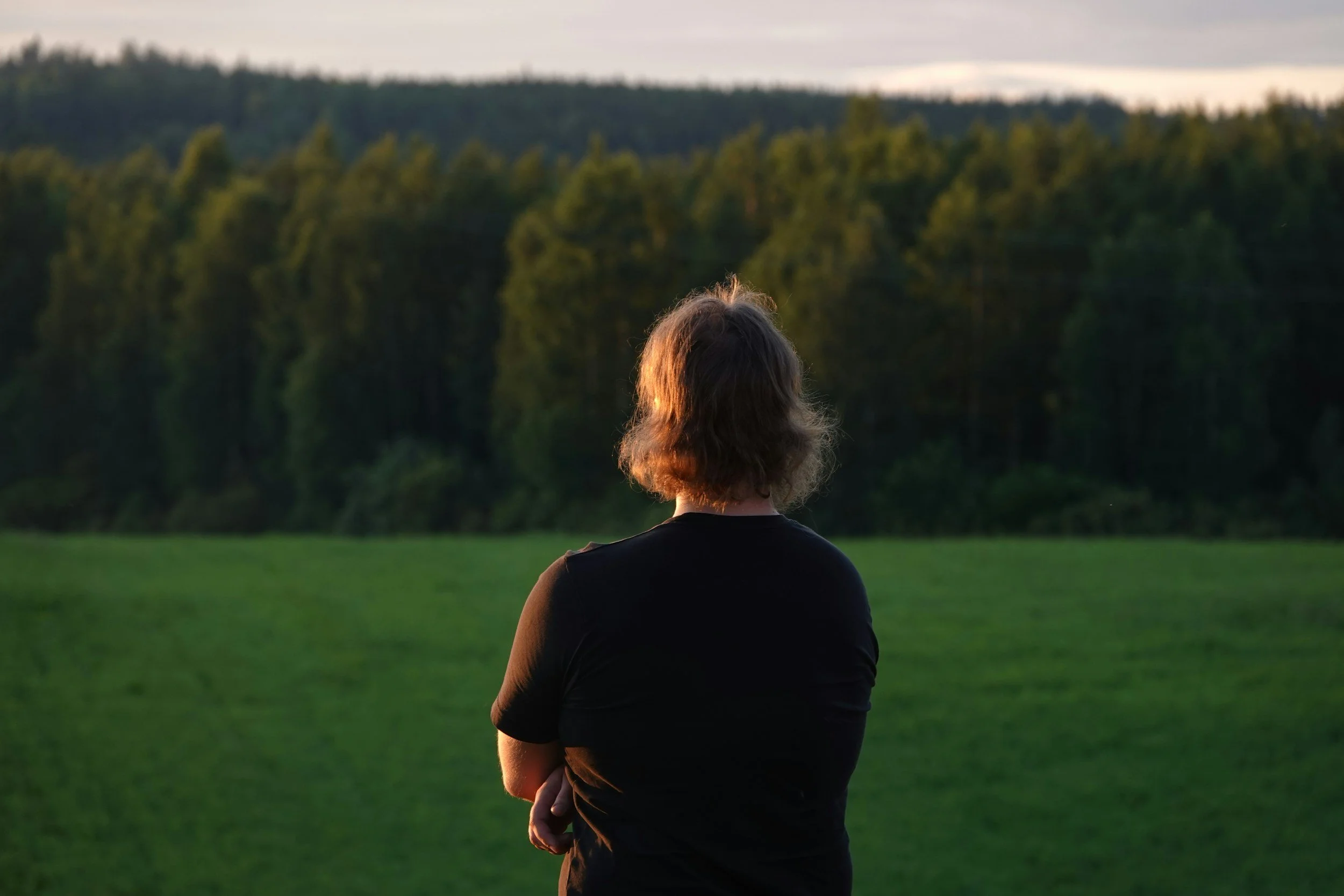 A person with shoulder-length hair wearing a black shirt, standing outdoors with arms crossed, looking at a distant forested landscape during sunset or dawn.
