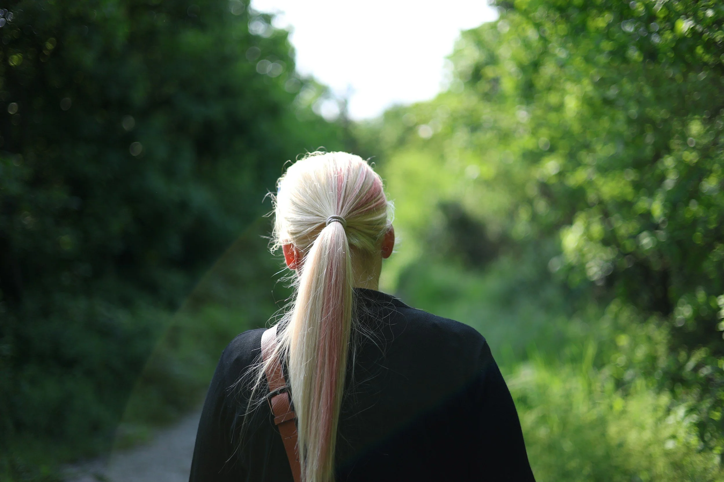 Back of a person with long blond hair tied in a ponytail walking on a forest trail surrounded by green trees and foliage