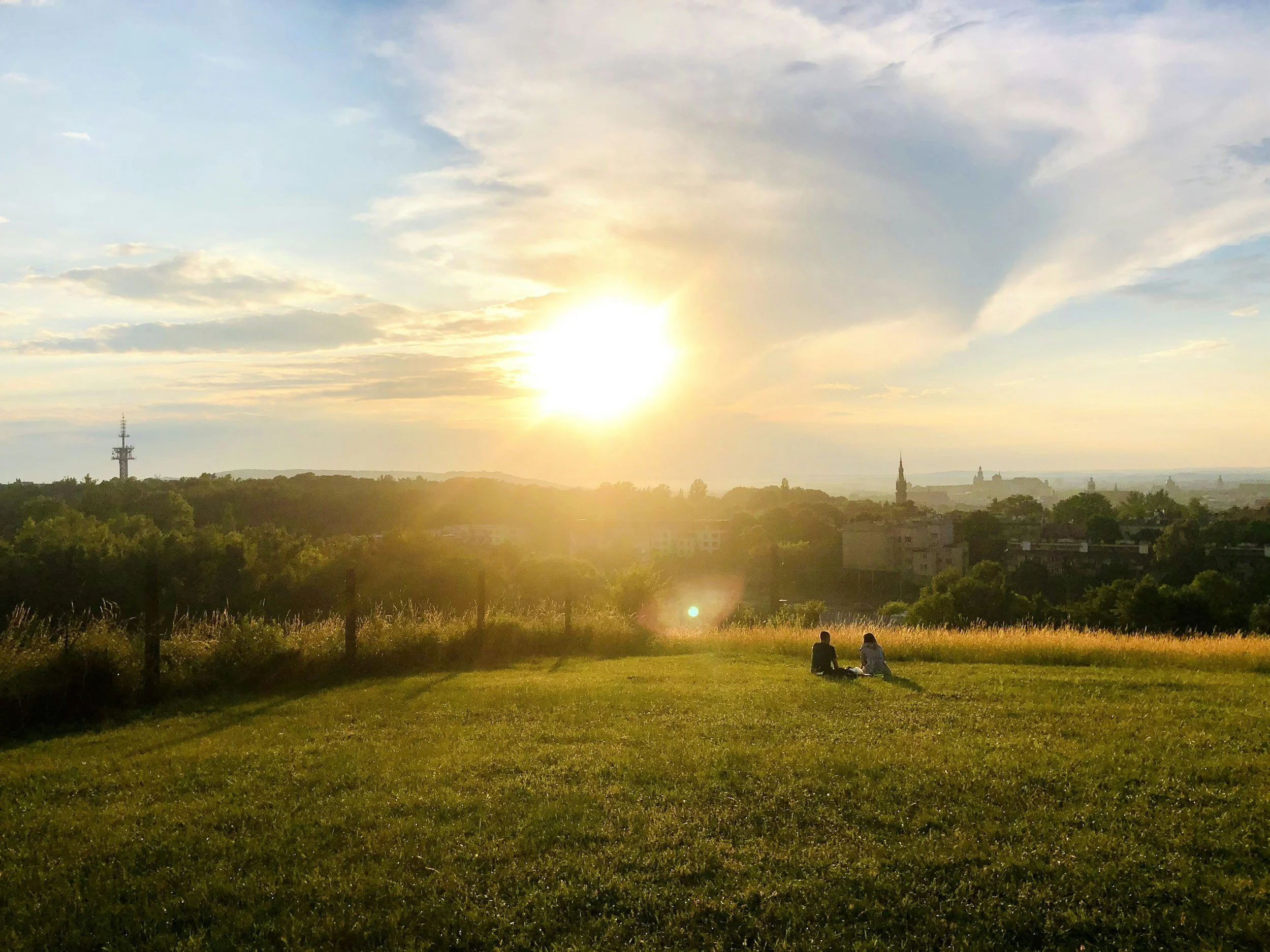 Sunset over a grassy park with two people sitting on the grass, trees, city skyline, and a tower in the distance.