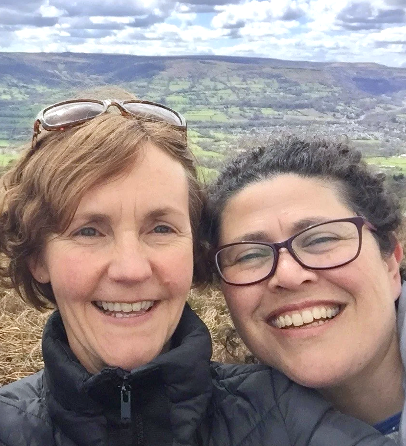 Two women smiling for a selfie outdoors with a scenic landscape of hills and fields in the background.