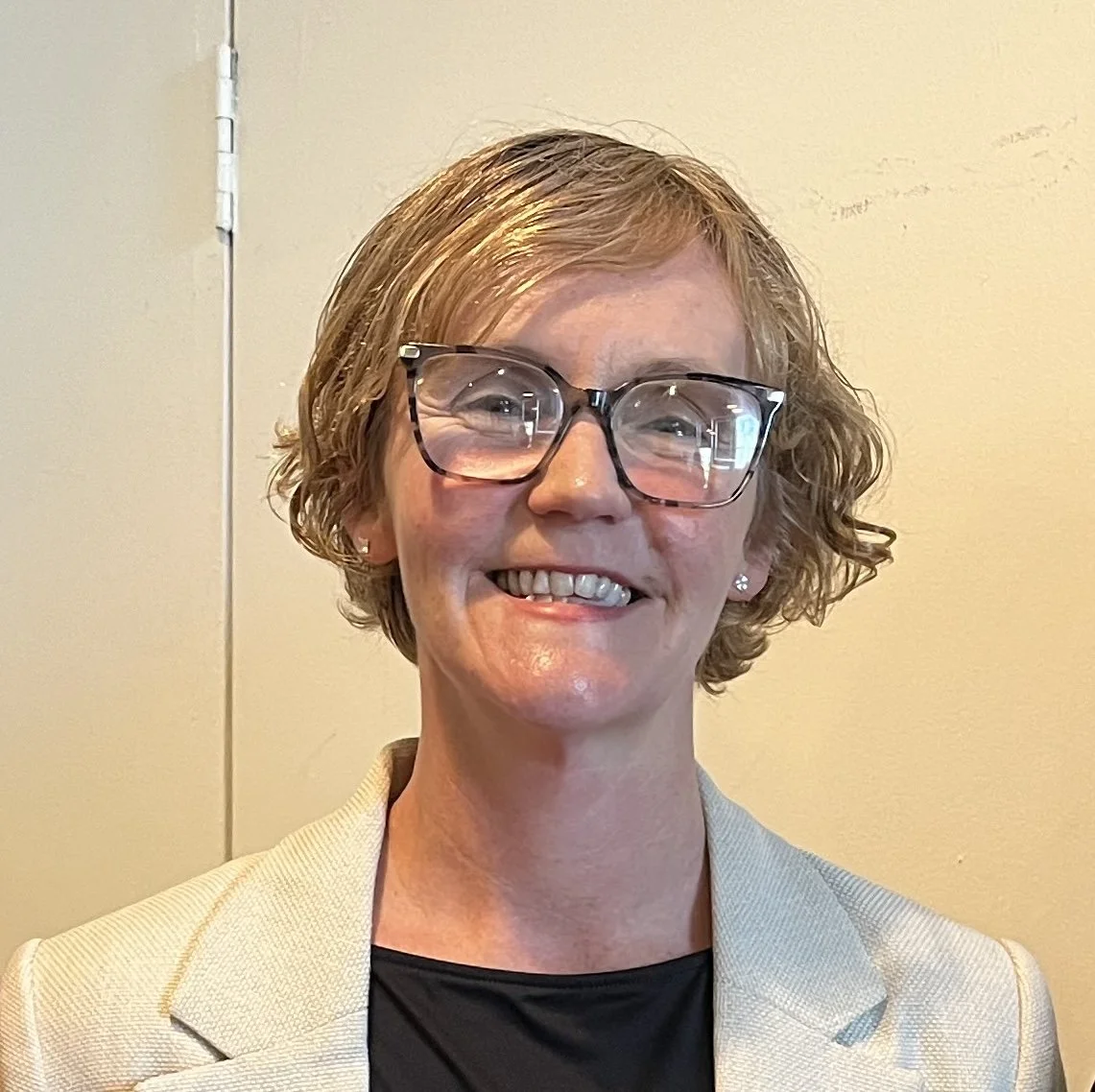 Close-up of a smiling woman wearing glasses and a beige blazer, standing against a plain yellow wall.