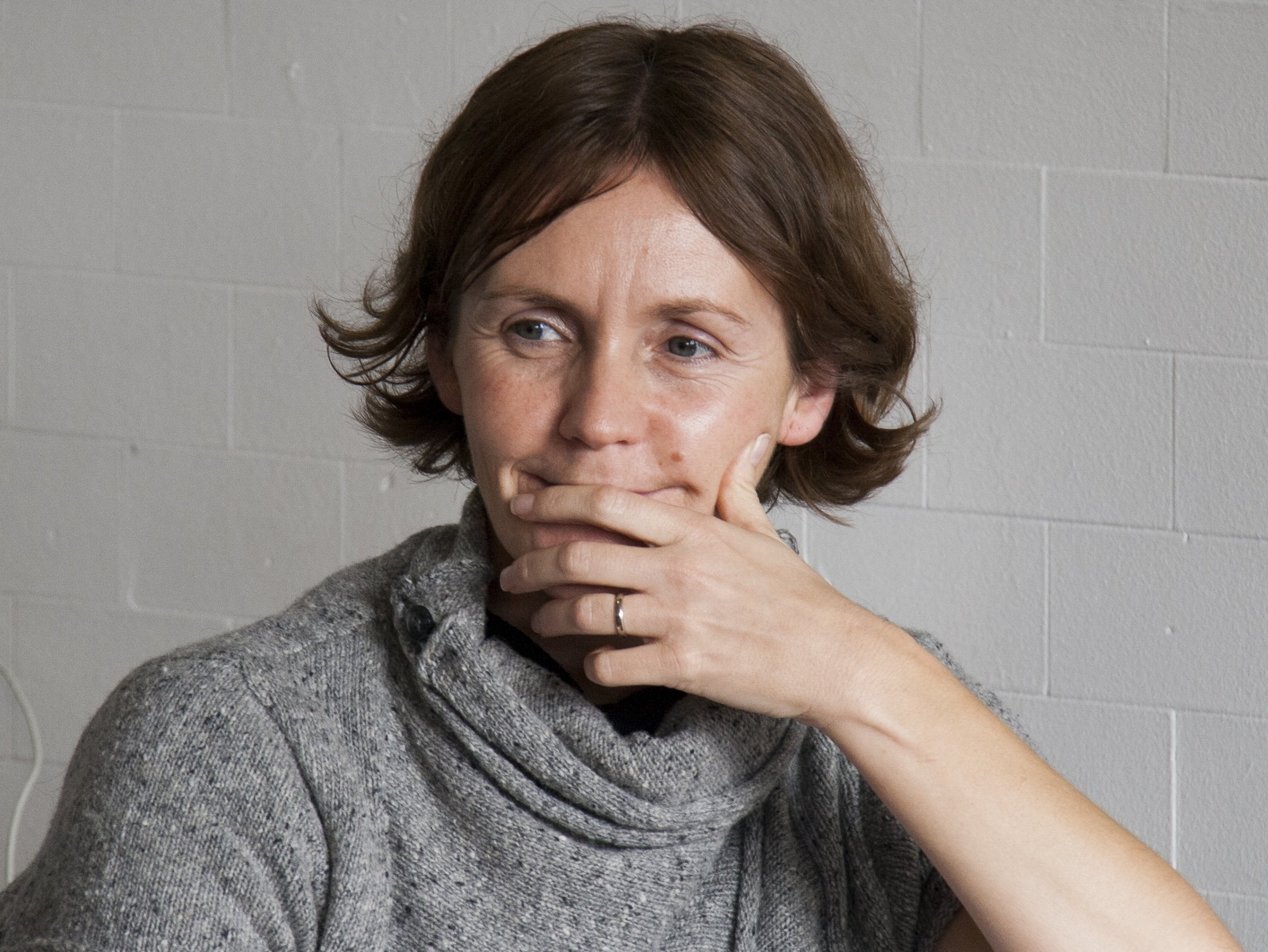 A woman with short brown hair and fair skin, wearing a gray sweater, is resting her chin on her hand in front of a plain gray wall.