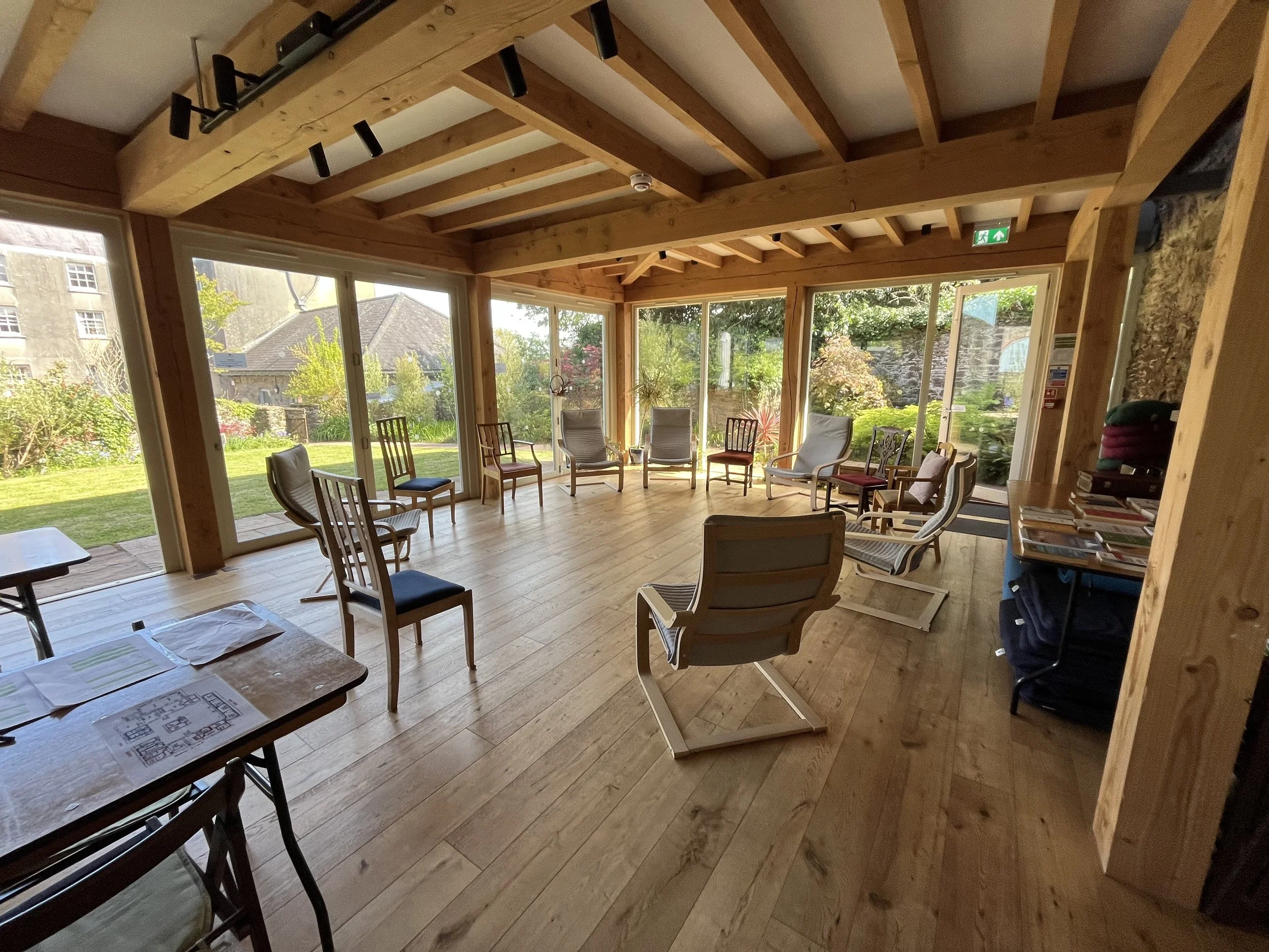 A bright sunroom with wooden beams on the ceiling and large windows showing a garden with trees, bushes, and a stone building. Inside, there are chairs arranged in a circle, a table with papers, and a bookshelf with books.